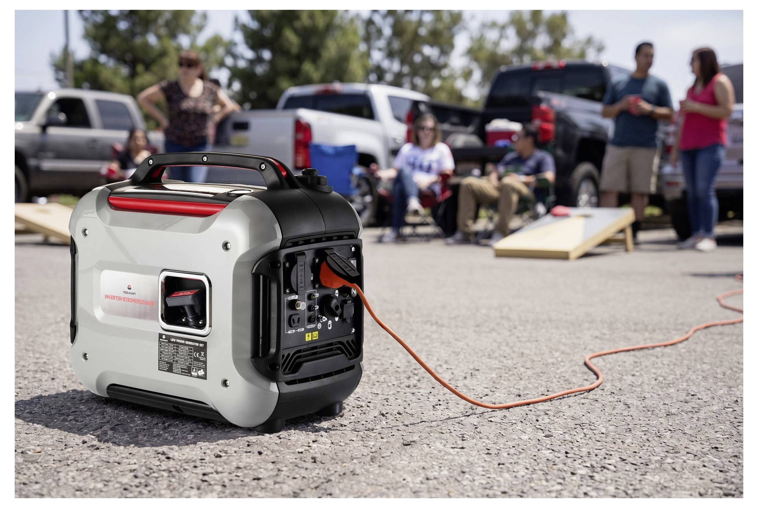 Ein tragbarer Generator auf einem Parkplatz versorgt ein Gerät mit Kabel mit Strom, während Menschen im Hintergrund zusammenkommen und Cornhole spielen.