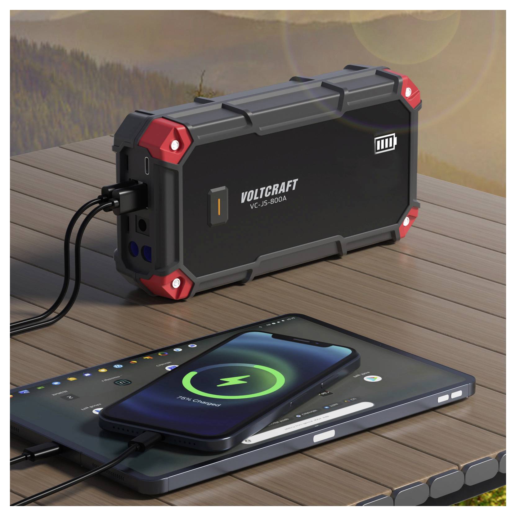 Tragbare Voltcraft-Stromstation, die ein Smartphone und ein Tablet auf einem Holztisch im Freien auflädt.