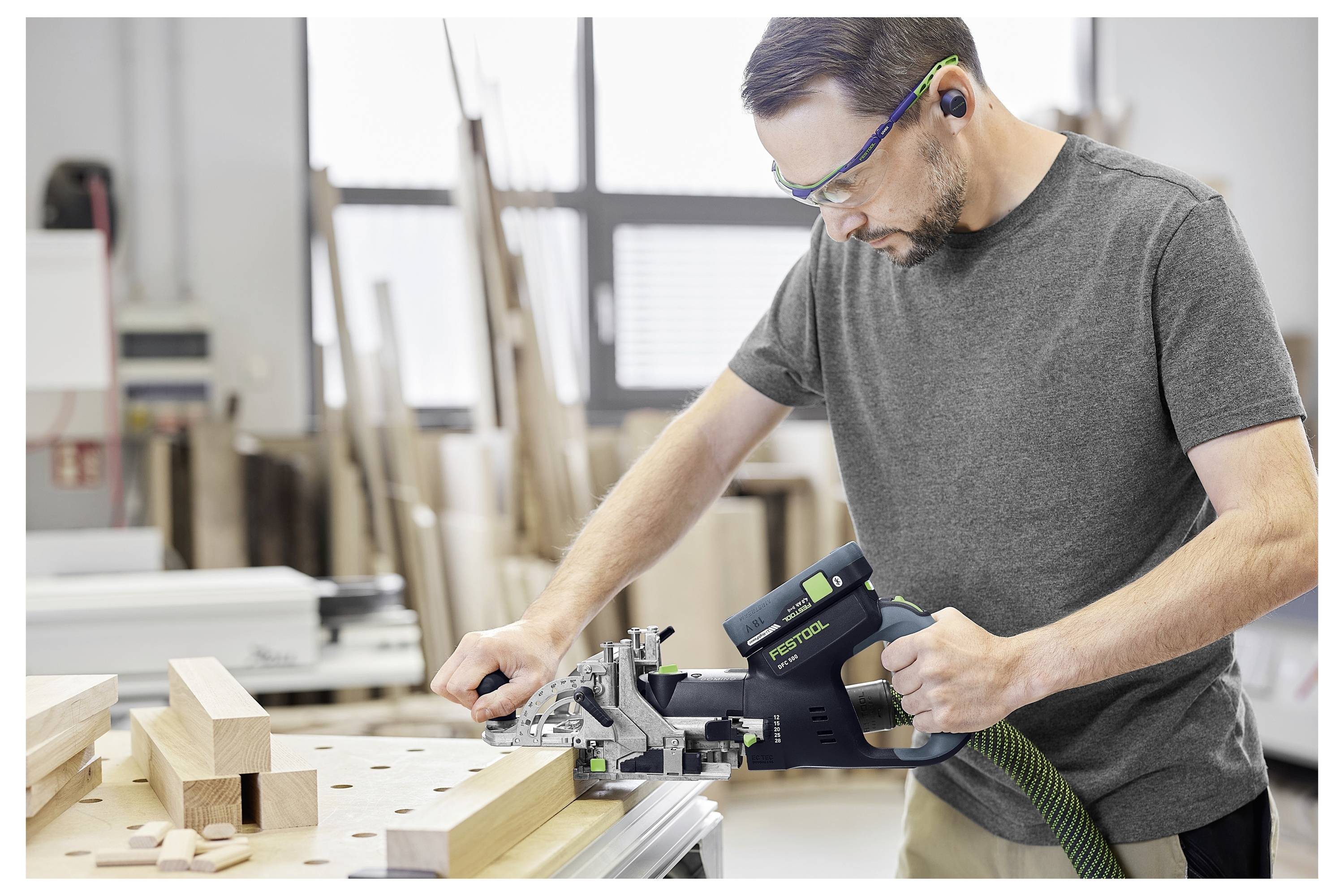 Ein Mann in Schutzbrille, der eine Festool-Verbindungsmaschine auf einem Holzbrett in einer Werkstatt verwendet. Holzteile und Werkzeuge sind im Hintergrund sichtbar.