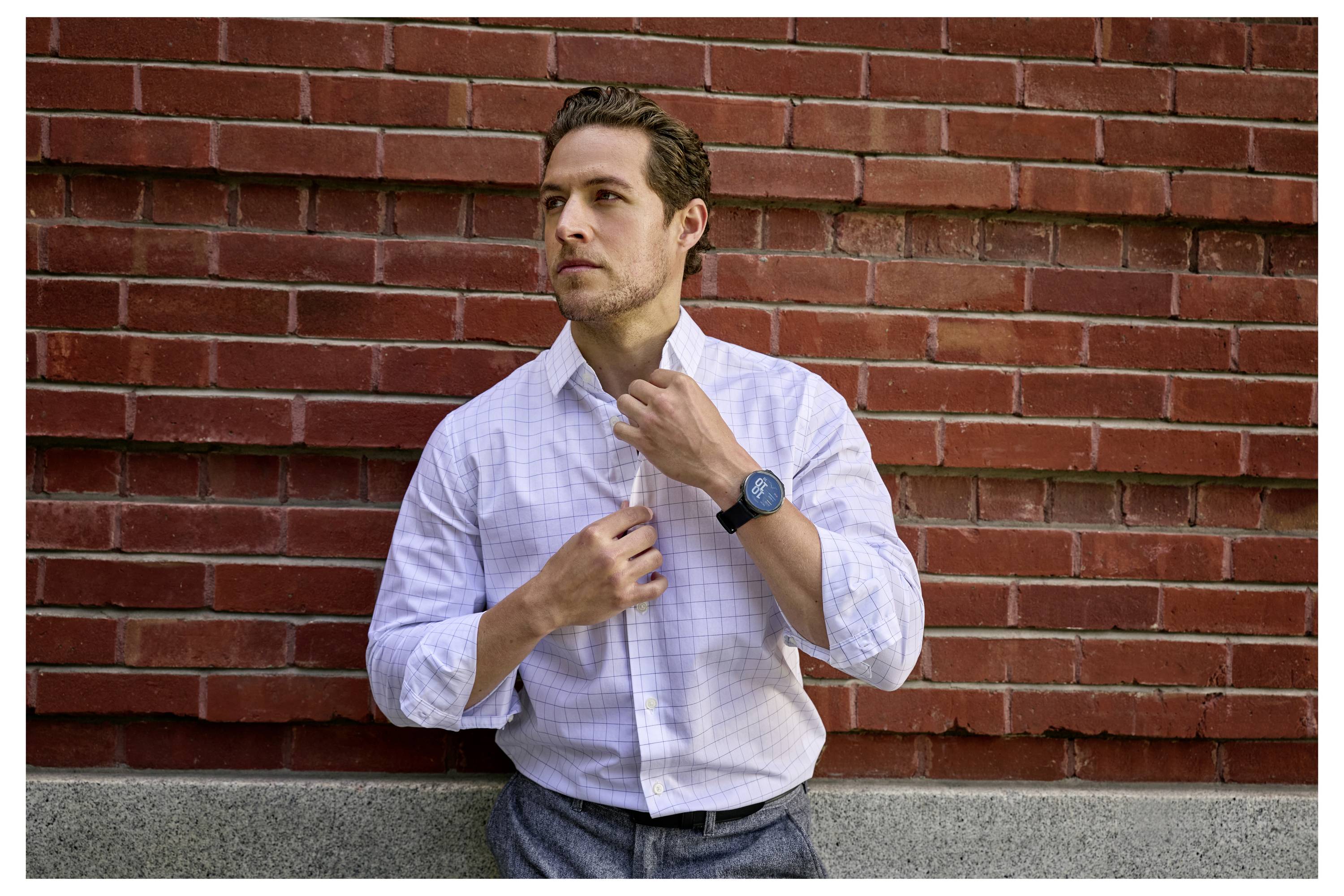 Eine Person stellt die Kragen ihres weißen Hemdes zurecht, während sie vor einer roten Ziegelmauer steht und eine dunkle Armbanduhr trägt.