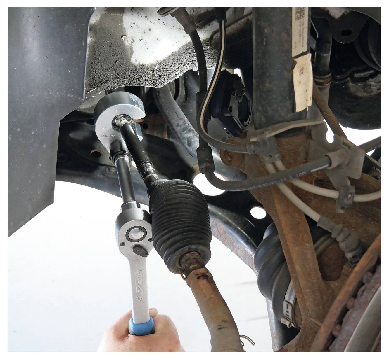 Eine Hand benutzt einen Schraubenschlüssel, um eine Schraube an der Unterseite eines Fahrzeugs einzustellen, wobei der Fokus auf den Aufhängungs- und Lenksystemen liegt.