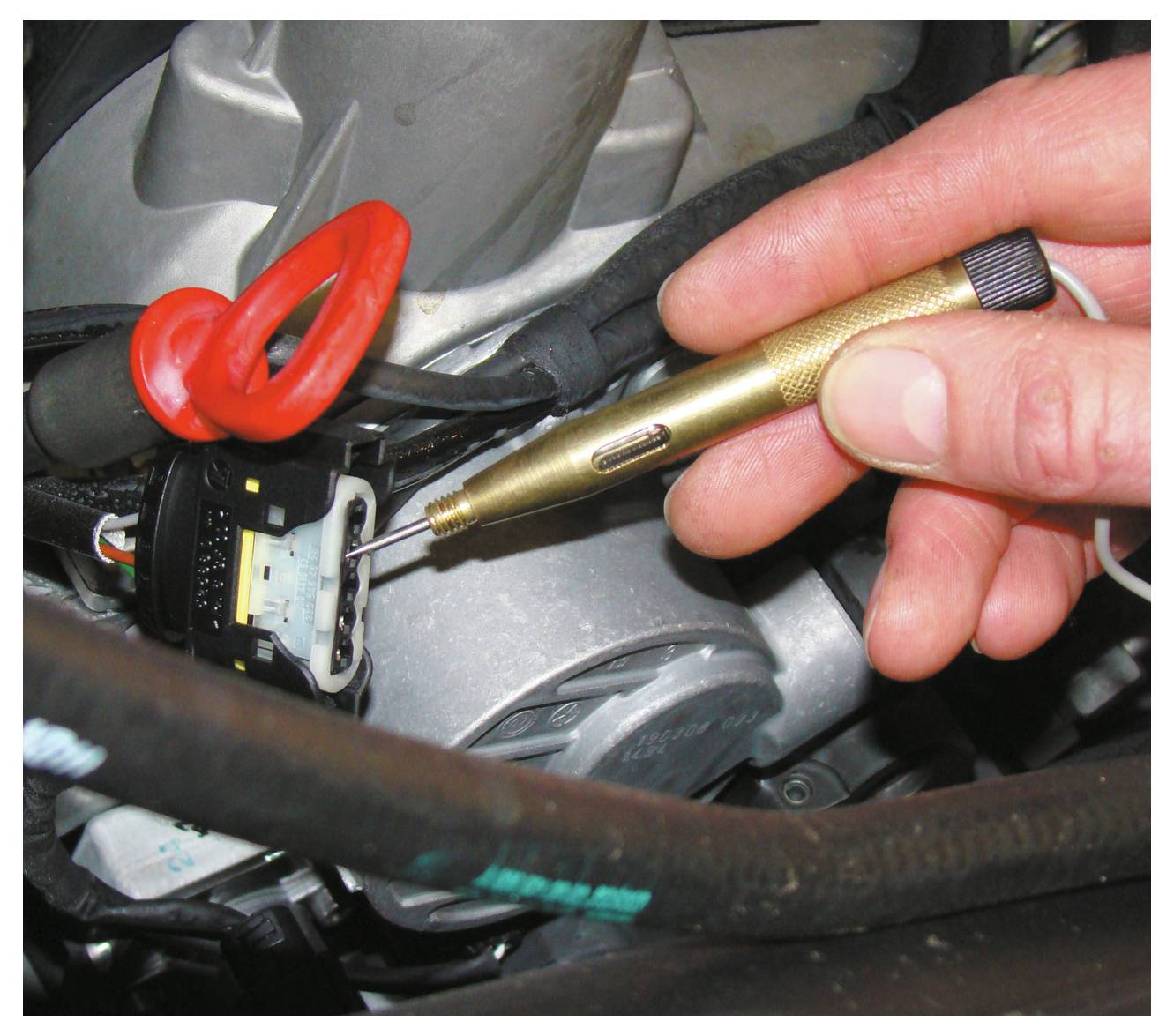 Eine Hand benutzt einen Spannungsprüfer, um einen elektrischen Stecker in einem Auto zu überprüfen. Die Sonde berührt einen Stift, wobei Drähte im Hintergrund sichtbar sind.