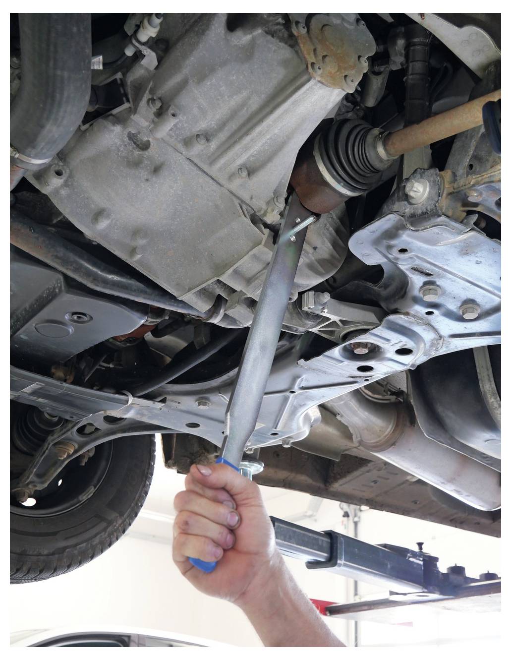 Ein Mechaniker verwendet einen Schraubenschlüssel, um eine Schraube auf der Unterseite eines Autos festzuziehen. Das Fahrzeug wird auf einer Hebebühne angehoben und zeigt Teile der Aufhängung und des Auspuffs.