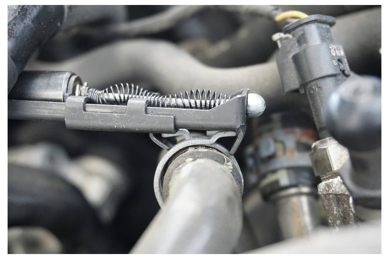 Eine Nahaufnahme eines Automotorkomponenten, das einen federbelasteten Spannmechanismus zeigt, umgeben von verschiedenen Schläuchen und Anschlüssen.