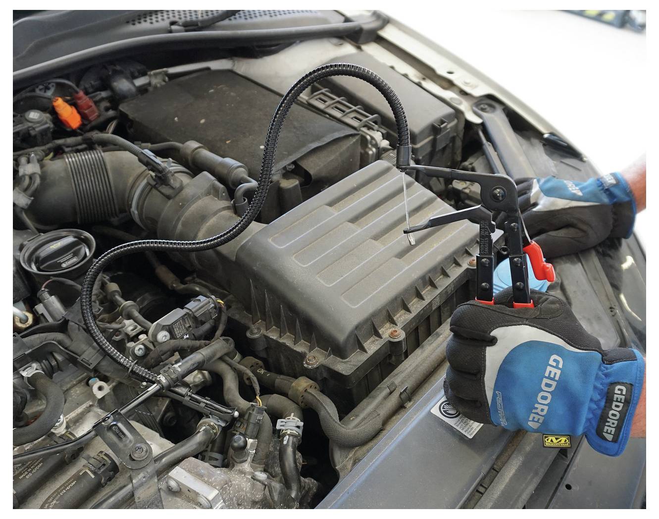 „Hände, die ein Werkzeug zur Überprüfung des Automotors verwenden; Nahaufnahme von Motorkomponenten und Werkzeug mit flexiblem Kabel. Handschuhe zeigen die Marke „Gedore