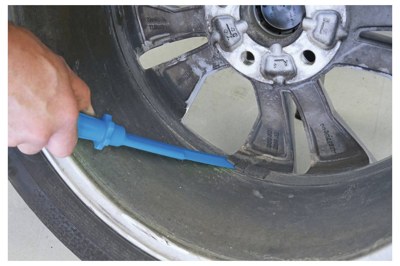 Eine Person hält ein blaues Hebelwerkzeug und inspiziert oder zeigt einen Schaden an der Felge eines Autoreifens, wobei sie sich auf den betroffenen Bereich in der Nähe der Reifenkante konzentriert.