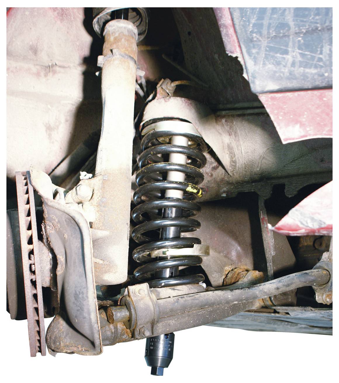 Ein Aufhängungssystem für ein Auto mit einer Schraubenfeder und einem Stoßdämpfer. Die Konstruktion ist mit dem Fahrgestell verbunden und zeigt Verschleiß- und Rostspuren.
