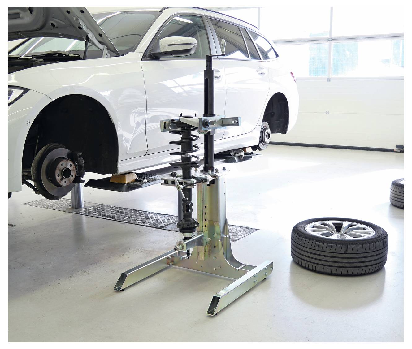 Ein weißes Auto steht erhöht auf einer Hebebühne in einer Garage. Sein Vorderrad ist entfernt. In der Nähe steht ein Reifenwechsler bereit, mit einem Ersatzreifen auf dem Boden.