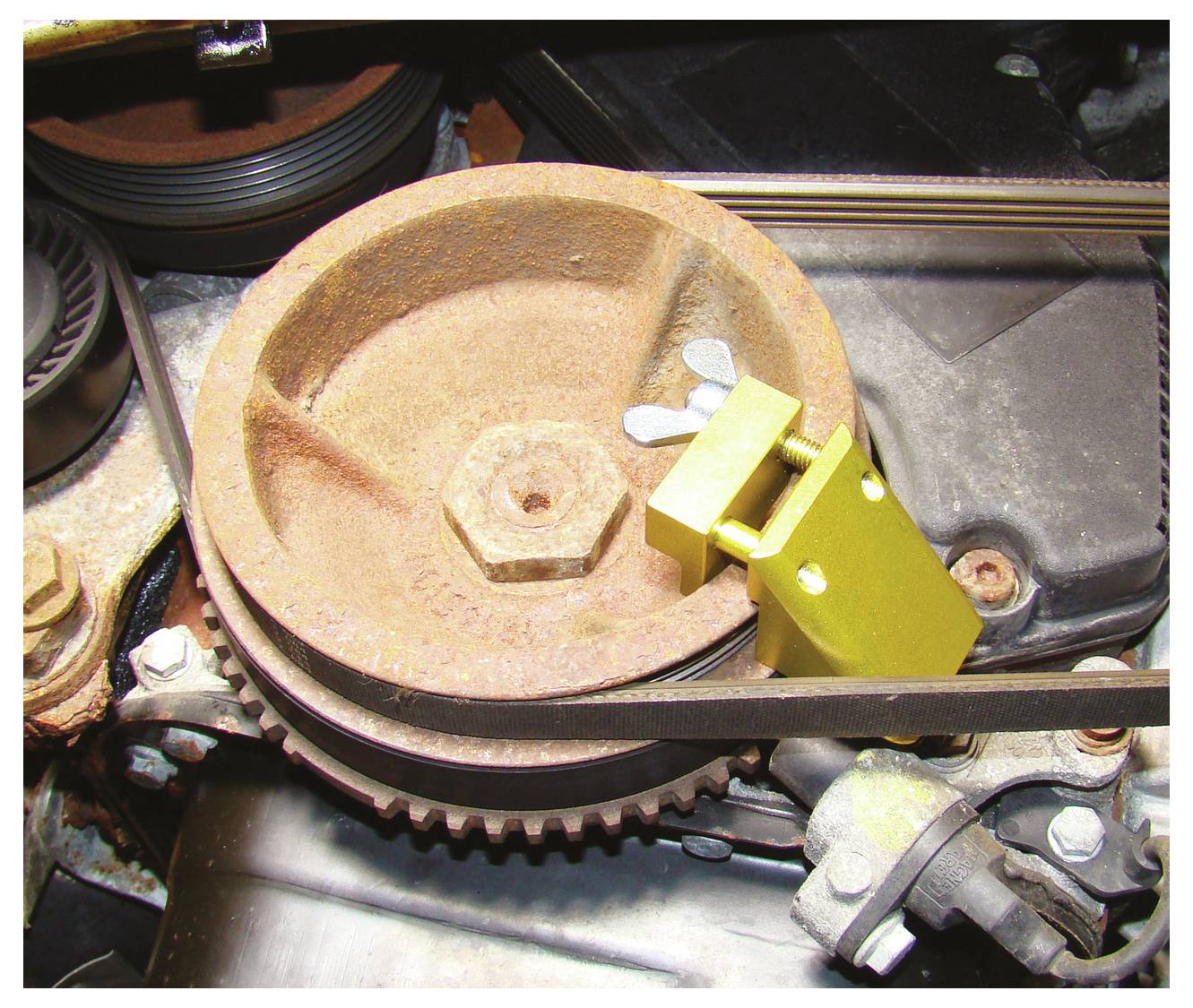 Ein Automotor mit einer verrosteten Riemenscheibe, gelbem Spannwerkzeug zur Wartung oder Einstellung befestigt, und einem sichtbaren Riemen auf der Riemenscheibe.