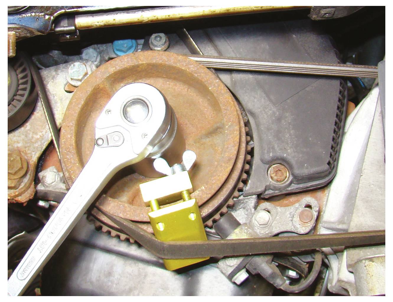 „Nahaufnahme eines Automotor-Riemenspanners mit einem Ratschenschlüssel und gelbem Spannersperrbolzen an Ort und Stelle, wobei mechanische Komponenten sichtbar sind.