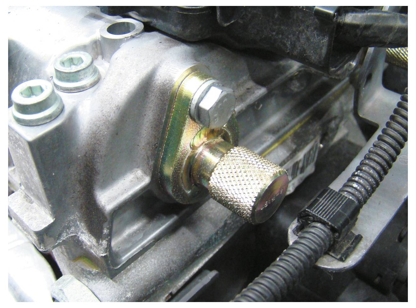 Nahaufnahme einer metallischen Ventilbaugruppe mit strukturiertem Knopf und umgebenden Motorteilen, die ihre Position und mechanischen Verbindungen hervorhebt.