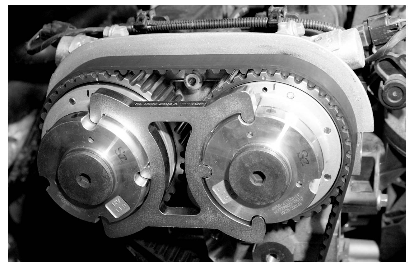 Schwarz-Weiß-Nahaufnahme eines Motorsteuersystems, das zwei Nockenwellen-Zahnräder und einen Zahnriemen zeigt und mechanische Präzision hervorhebt.