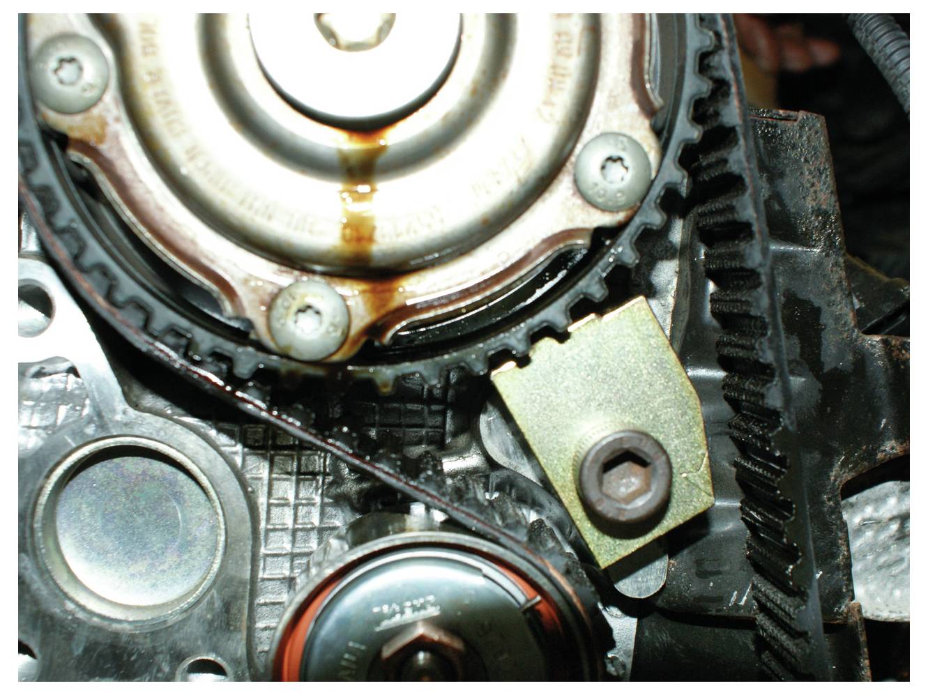 Nahaufnahme des Zahnriemensystems eines Automotors mit einer Spannrolle, einer Metallhalterung und sichtbaren Ölrückständen zur Wartungsreferenz.