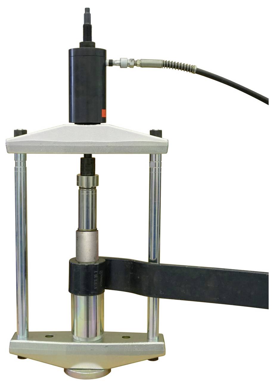 Eine hydraulische Presse mit einem schwarzen Kolben, verbunden durch Kabel, mit zwei vertikalen Stangen und einem Metallsockel, verwendet zum Komprimieren von Materialien.