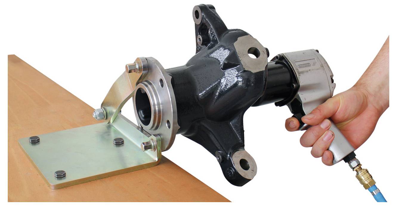 Eine Hand bedient ein Werkzeug, um ein großes schwarzes mechanisches Bauteil auf einer Metallplatte zu befestigen, die auf einer Holzoberfläche angebracht ist.