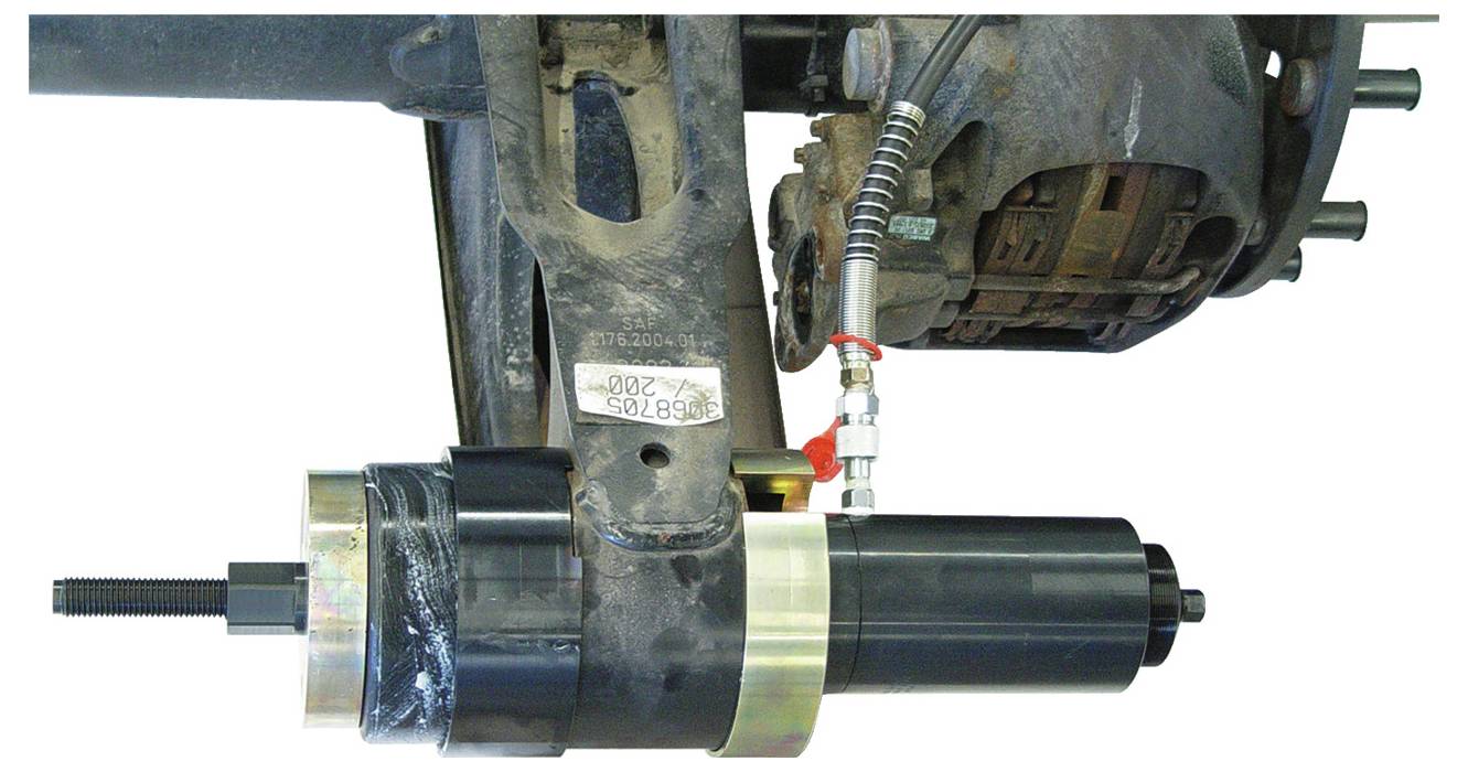 Ein hydraulisches Werkzeug ist an einer Aufhängungskomponente eines Fahrzeugs angebracht, wahrscheinlich für Wartungs- oder Reparaturzwecke, wobei Verbindungen und Armaturen sichtbar sind.