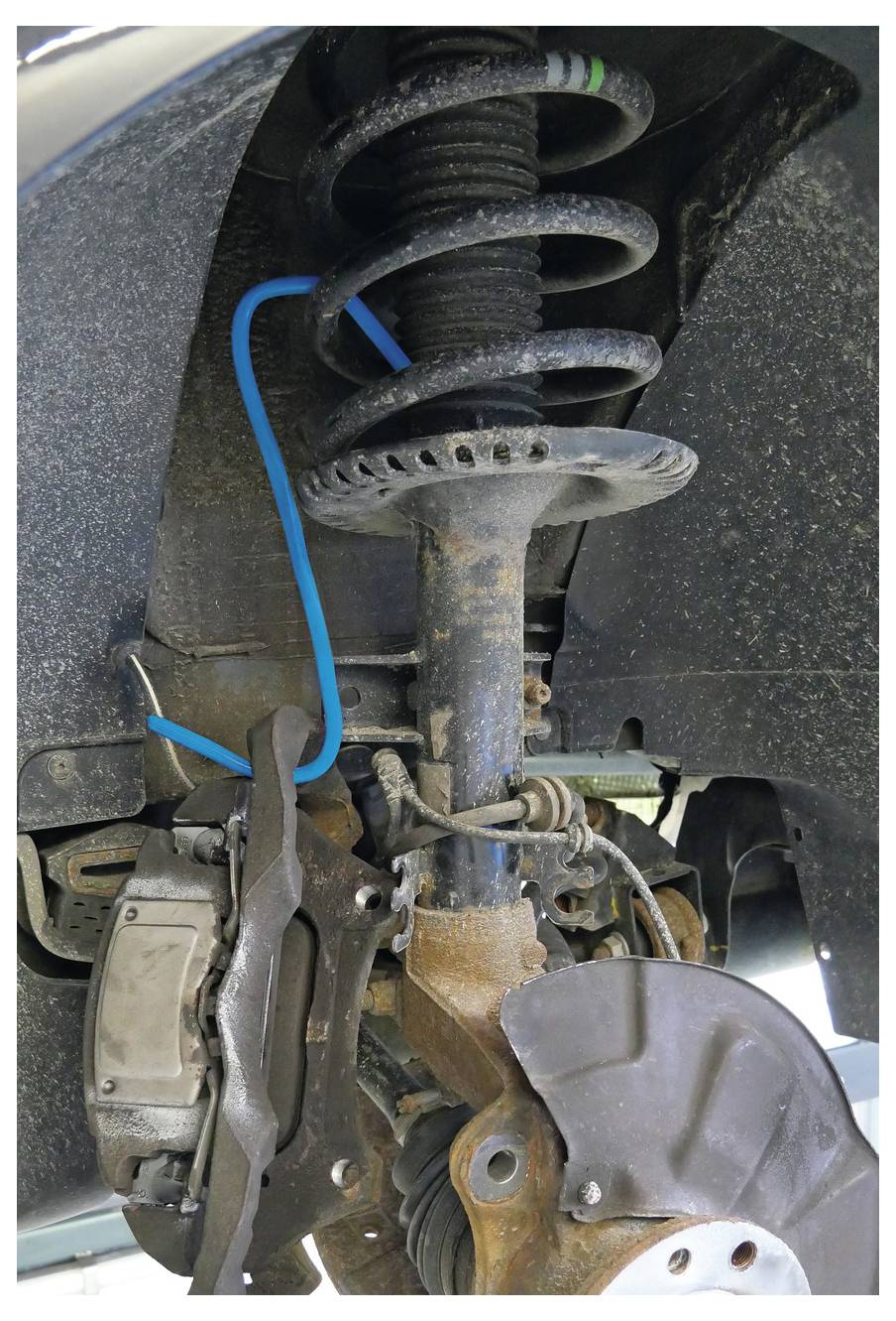 Nahaufnahme des Aufhängungssystems eines Autos mit einer Schraubenfeder, einem Stoßdämpfer und Bremsenkomponenten, die Verschleiß und Rost zeigen.