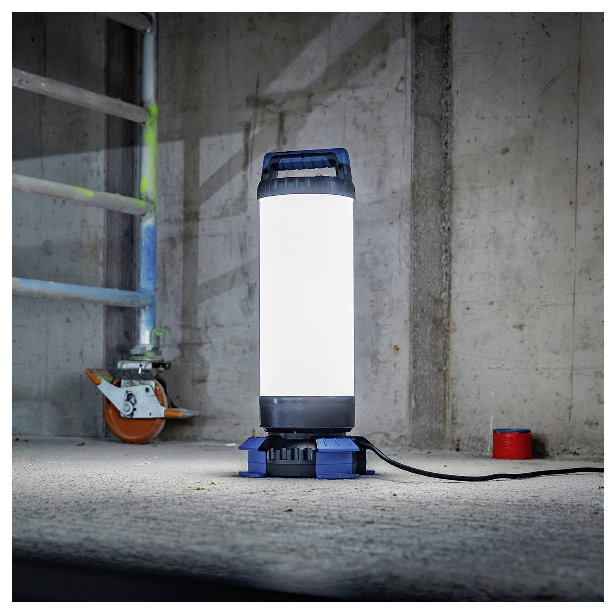 Ein helles Arbeitslicht auf einem grauen Betonboden neben einer Leiter und einem roten Objekt, das einen schwach beleuchteten Industrieraum erhellt.