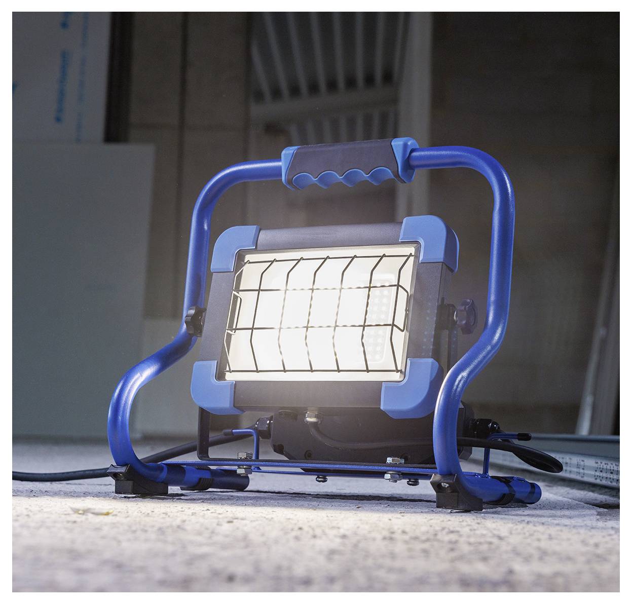 Eine blaue tragbare Baustrahler beleuchtet einen Betonboden in einem schwach beleuchteten Raum und unterstreicht damit dessen Robustheit und industrielles Design.