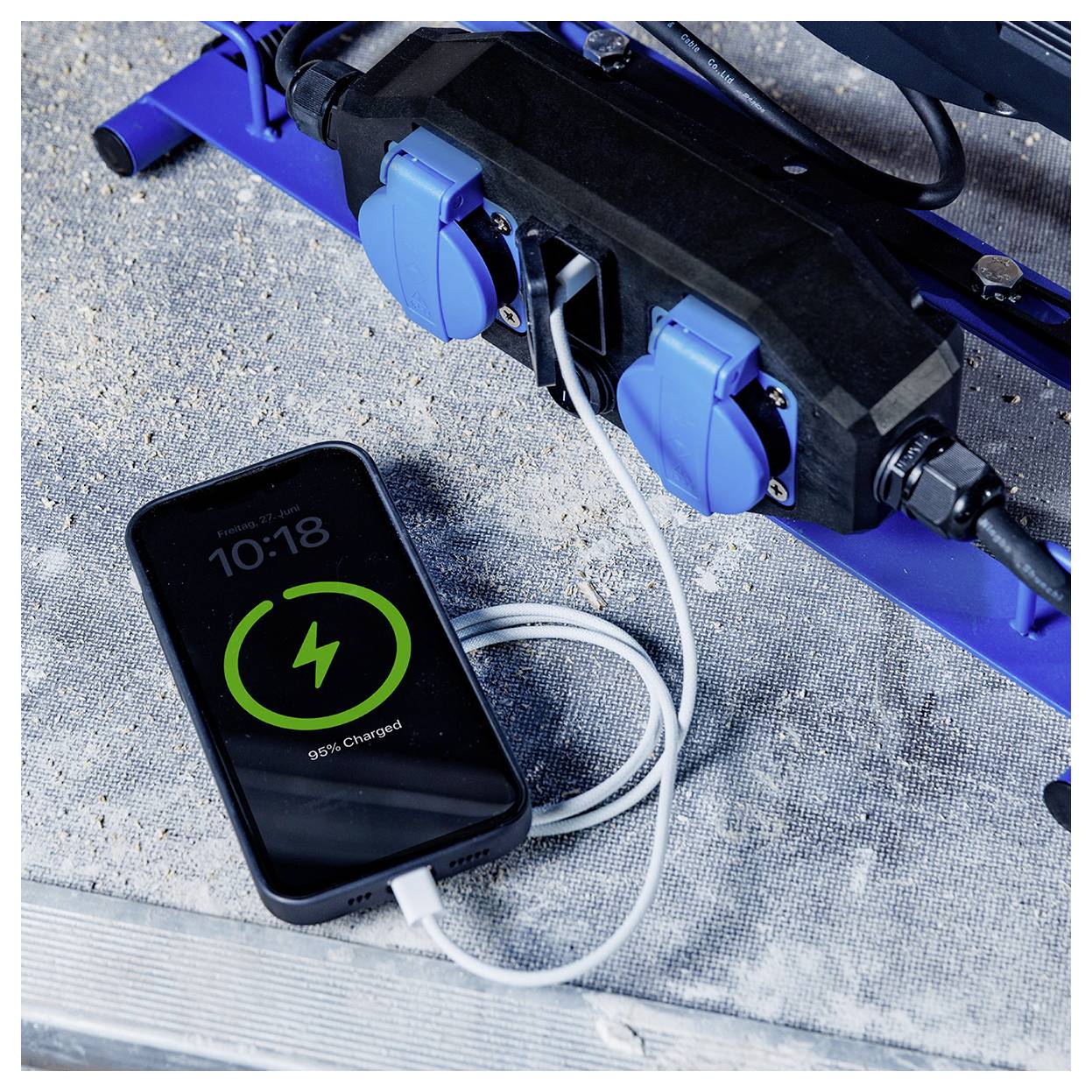 Ein Smartphone mit 95% Ladung, das an eine Steckdosenleiste mit mehreren Steckplätzen auf einer Metalloberfläche angeschlossen ist.