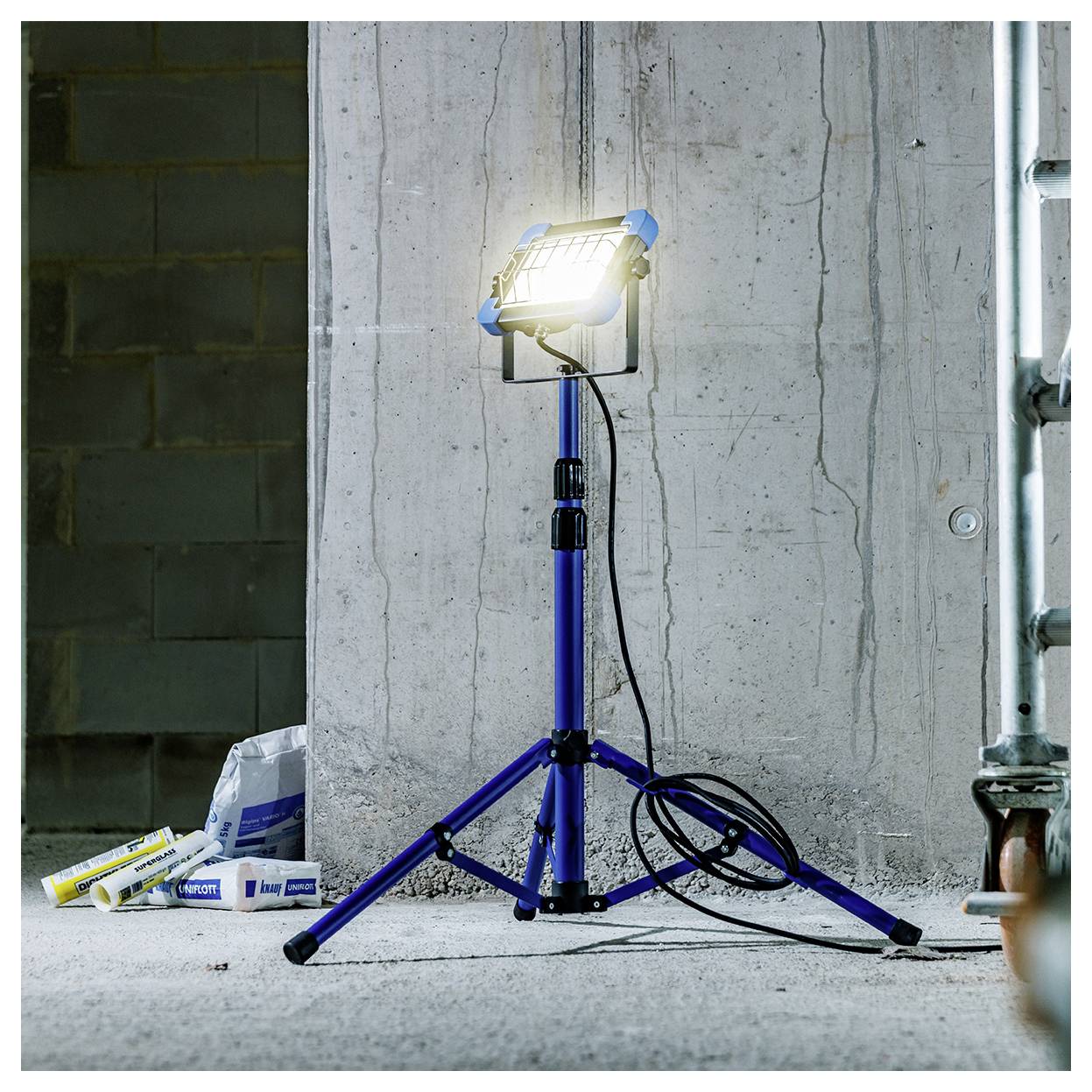 Ein helles Arbeitslicht auf einem blauen Stativ steht auf einem Betonboden und beleuchtet eine Baustelle mit Zementwänden und Baumaterialien.