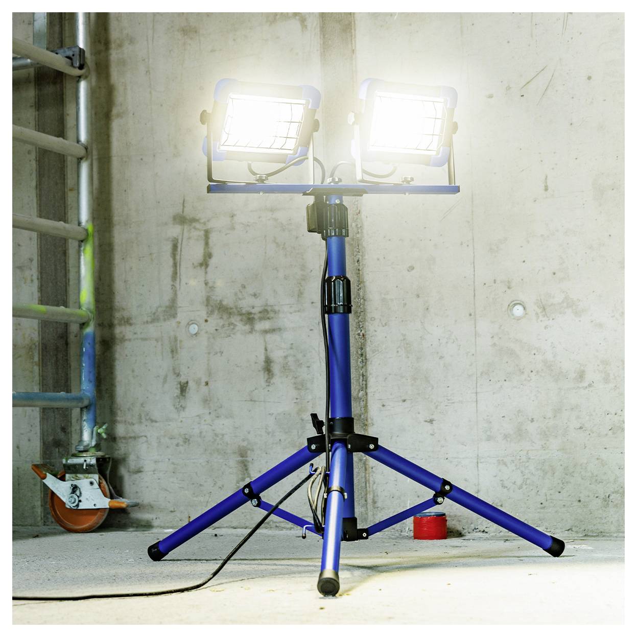 Eine tragbare Arbeitsleuchte mit doppeltem blauem Kopf auf einem Stativständer beleuchtet einen Betonbaustellen bereich.