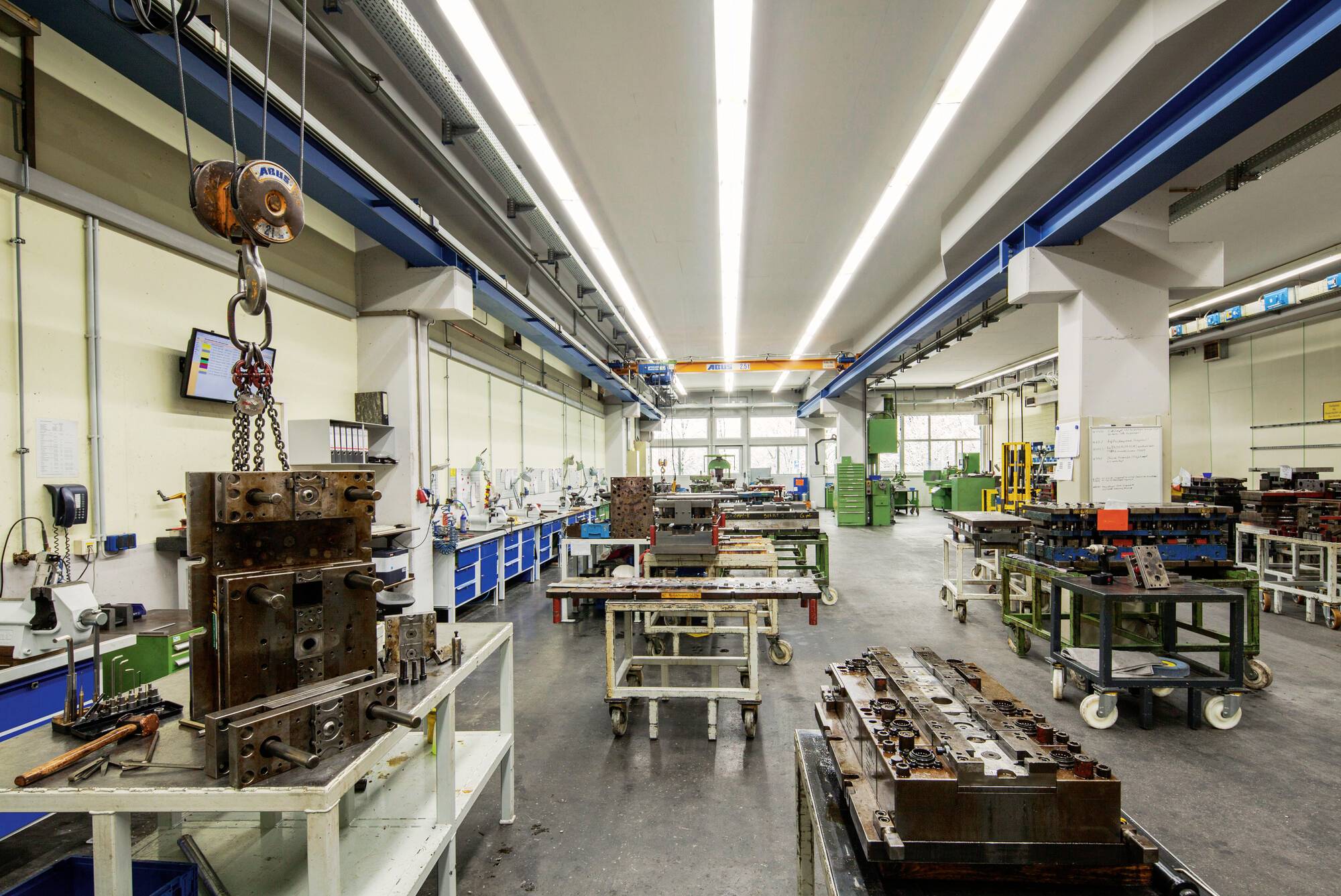 Inneres einer Metallwerkstatt mit Maschinen, Werkbänken und Laufkran. Werkzeuge und Ausrüstung sind an den Wänden und auf Tischen organisiert.