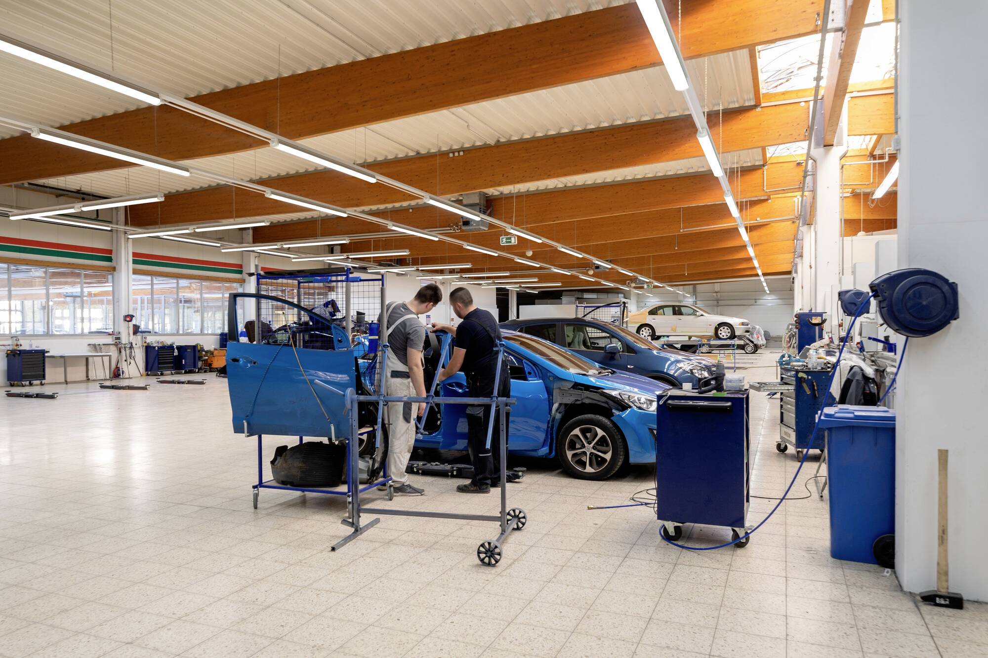 Zwei Mechaniker arbeiten in einer geräumigen, hell beleuchteten Werkstatt an einem Auto. Verschiedene Werkzeuge und Ausrüstungen umgeben sie und unterstreichen ein Kfz-Reparaturumfeld.