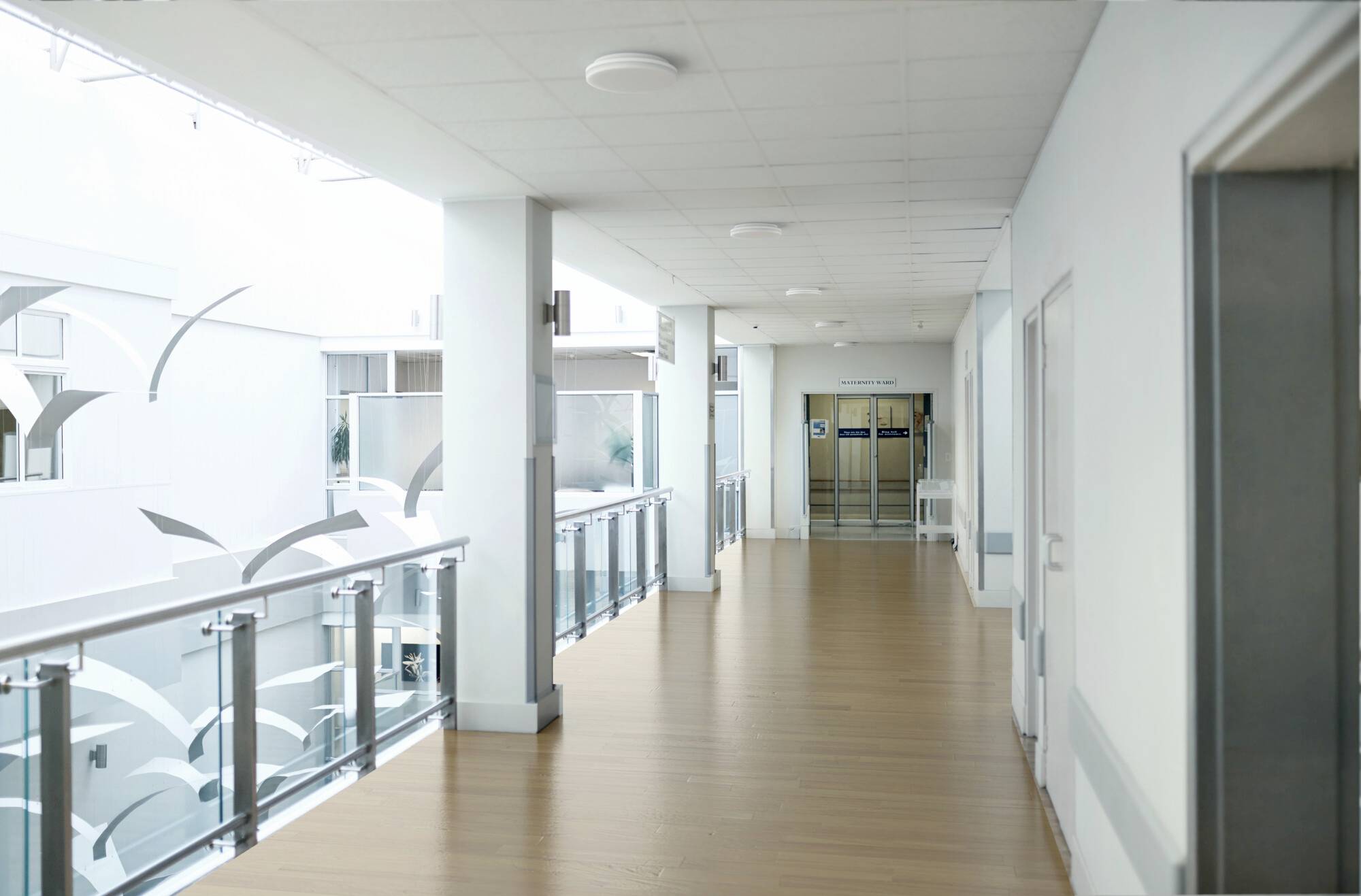 Ein Krankenhauskorridor mit Holzböden, Glasgeländern und einem Schild, das einen Aufzug am fernen Ende anzeigt.