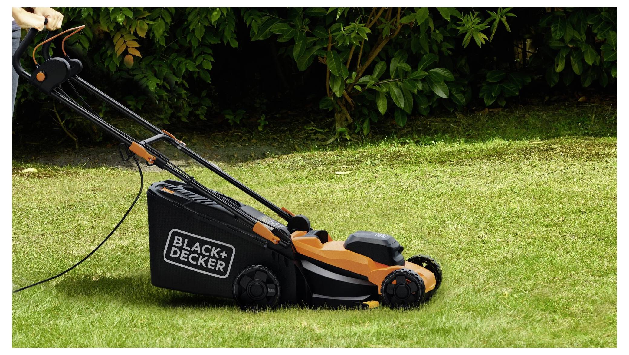 Eine Person schiebt einen Black+Decker Rasenmäher über einen grünen Rasen, mit Büschen im Hintergrund. Der Rasenmäher ist schwarz und orange.