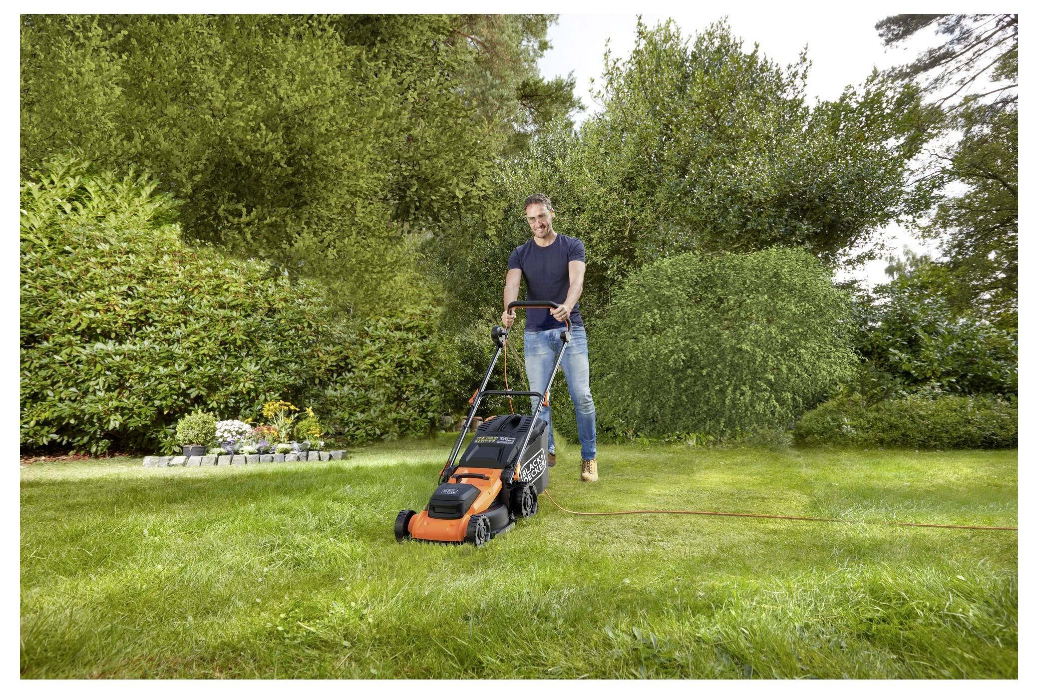 Eine Person mäht einen üppigen, grünen Rasen mit einem orange-schwarzen Rasenmäher. Im Hintergrund umgeben Bäume und Sträucher den Hof.