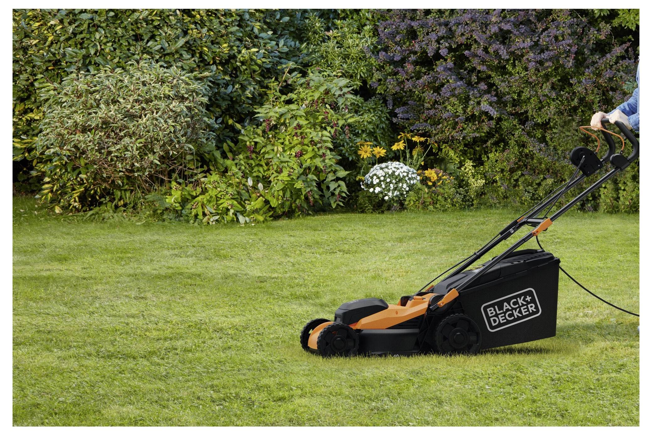 Eine Person benutzt einen Black+Decker-Rasenmäher auf einem ordentlich gestutzten Rasen, umgeben von üppigen, grünen Büschen und blühenden Pflanzen.