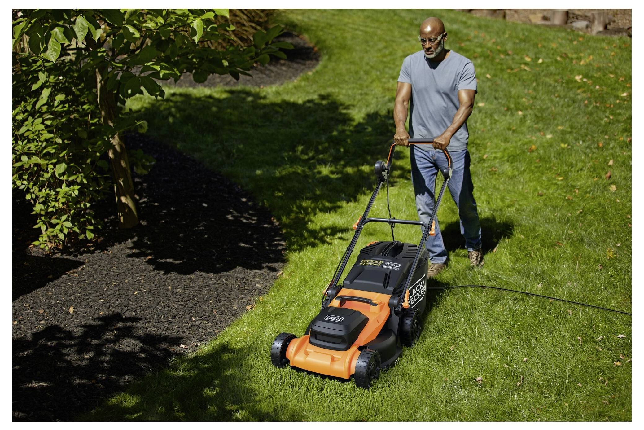 Eine Person, die einen lebendigen grünen Rasen mit einem orange-schwarzen Elektrorasenmäher mäht, umgeben von schattenspendenden Bäumen und Sträuchern.