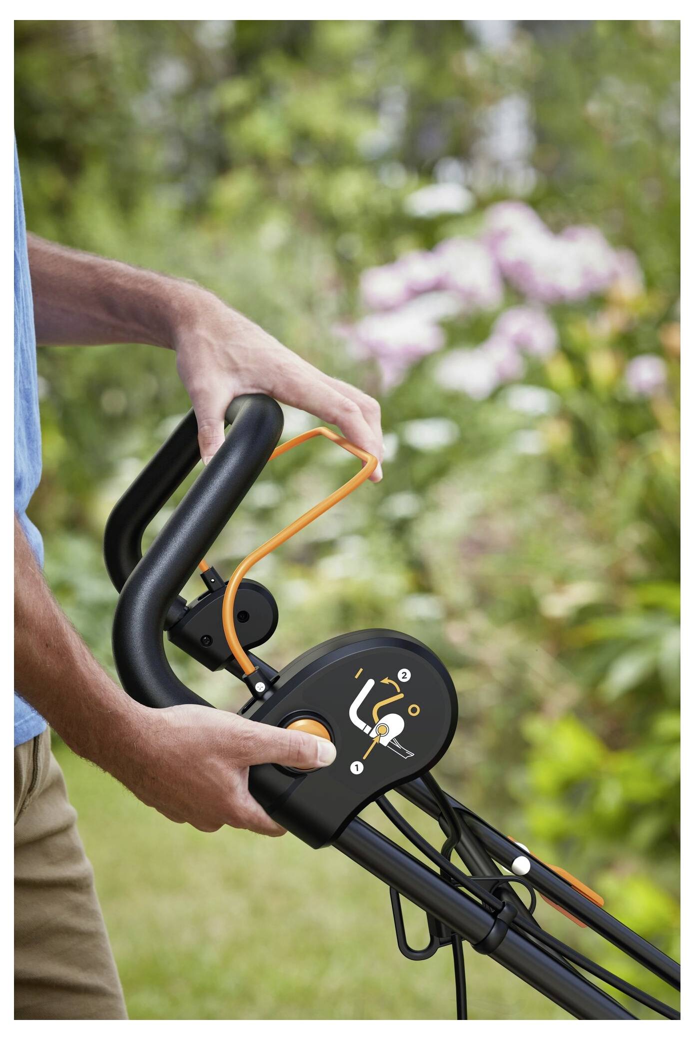 Person holding einen Rasenmäher-Griff mit Fokus auf den orangefarbenen Sicherheitshebel, umgeben von einem Garten mit blühenden Blumen.