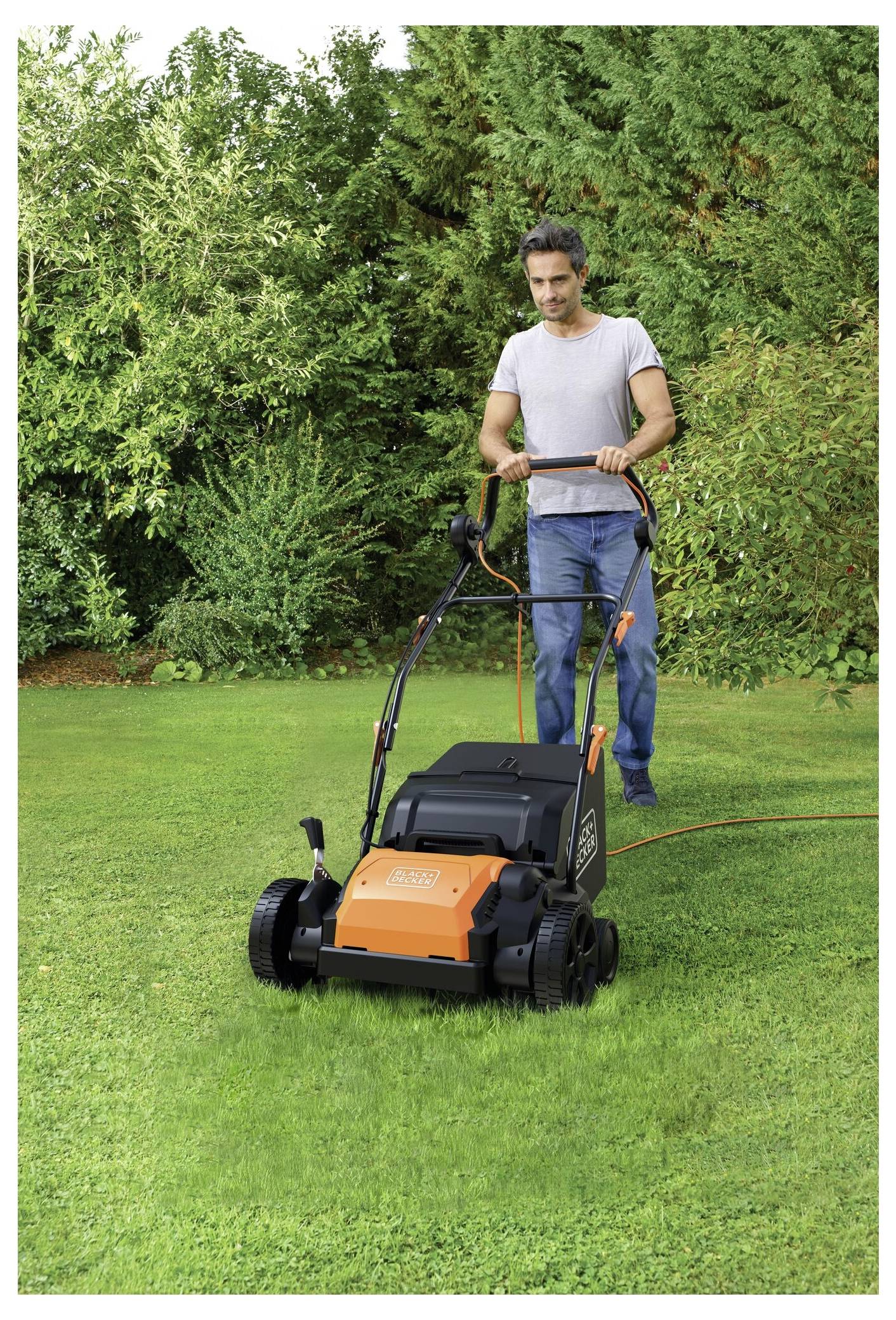 Eine Person benutzt einen Rasenbelüfter auf einem grünen Rasen, umgeben von üppigem Grün. Die Maschine hat ein orange-schwarzes Design.