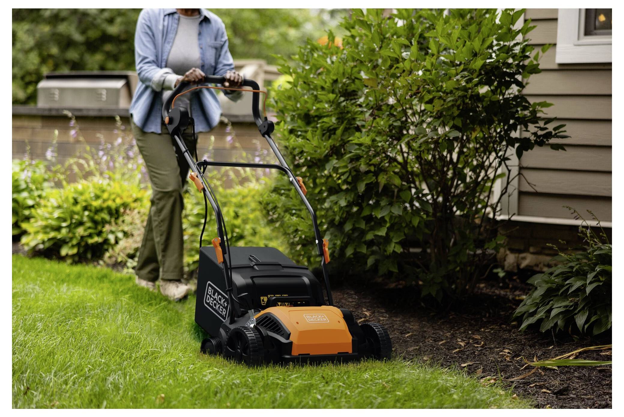 Eine Person mäht einen Rasen mit einem orange-schwarzen elektrischen Rasenmäher neben einem Blumenbeet an einer Hauswand.