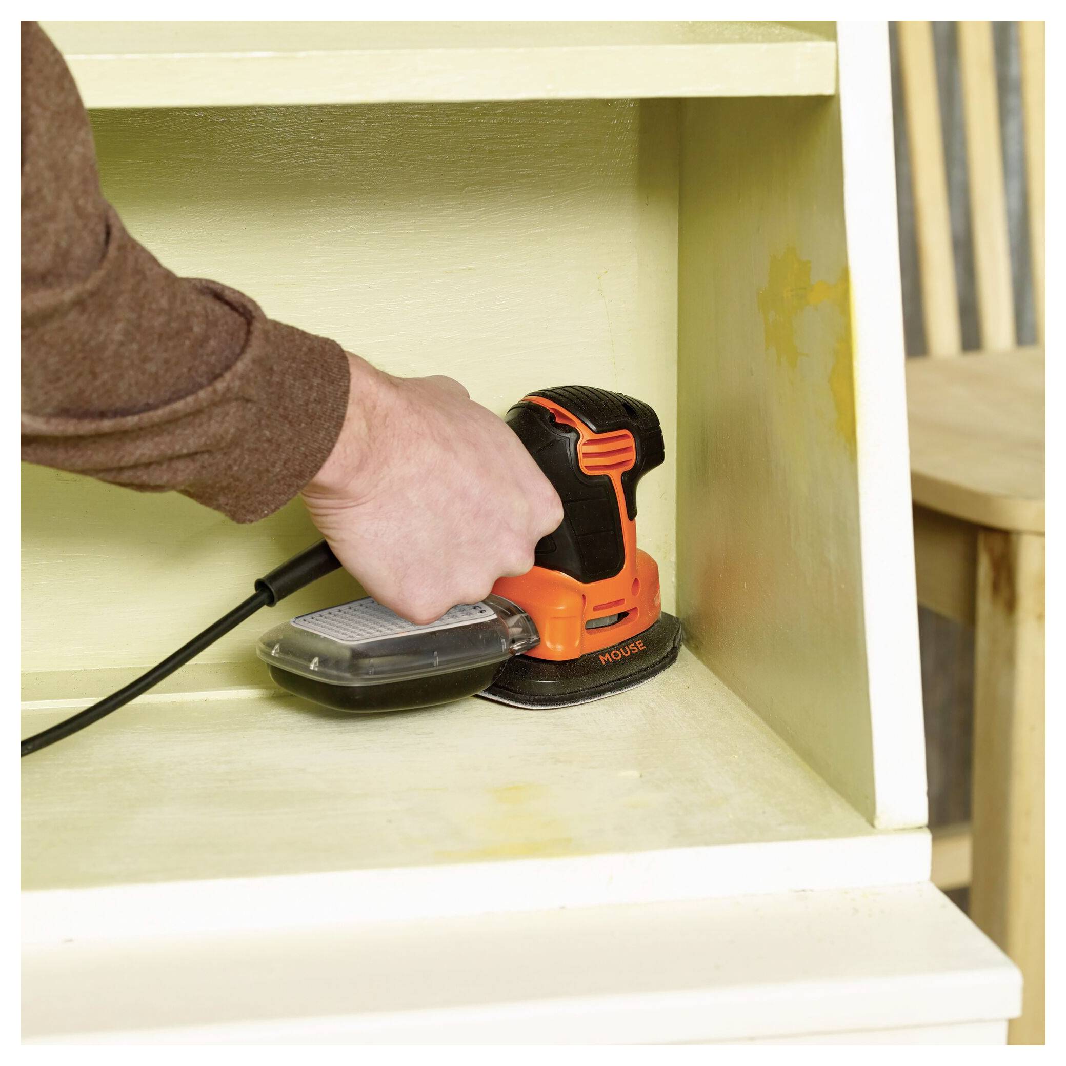 Eine Person benutzt einen elektrischen Schleiffer an einem Möbelstück aus Holz und glättet dessen Oberfläche. Der Schleiffer ist orange und schwarz und in eine Steckdose eingesteckt.