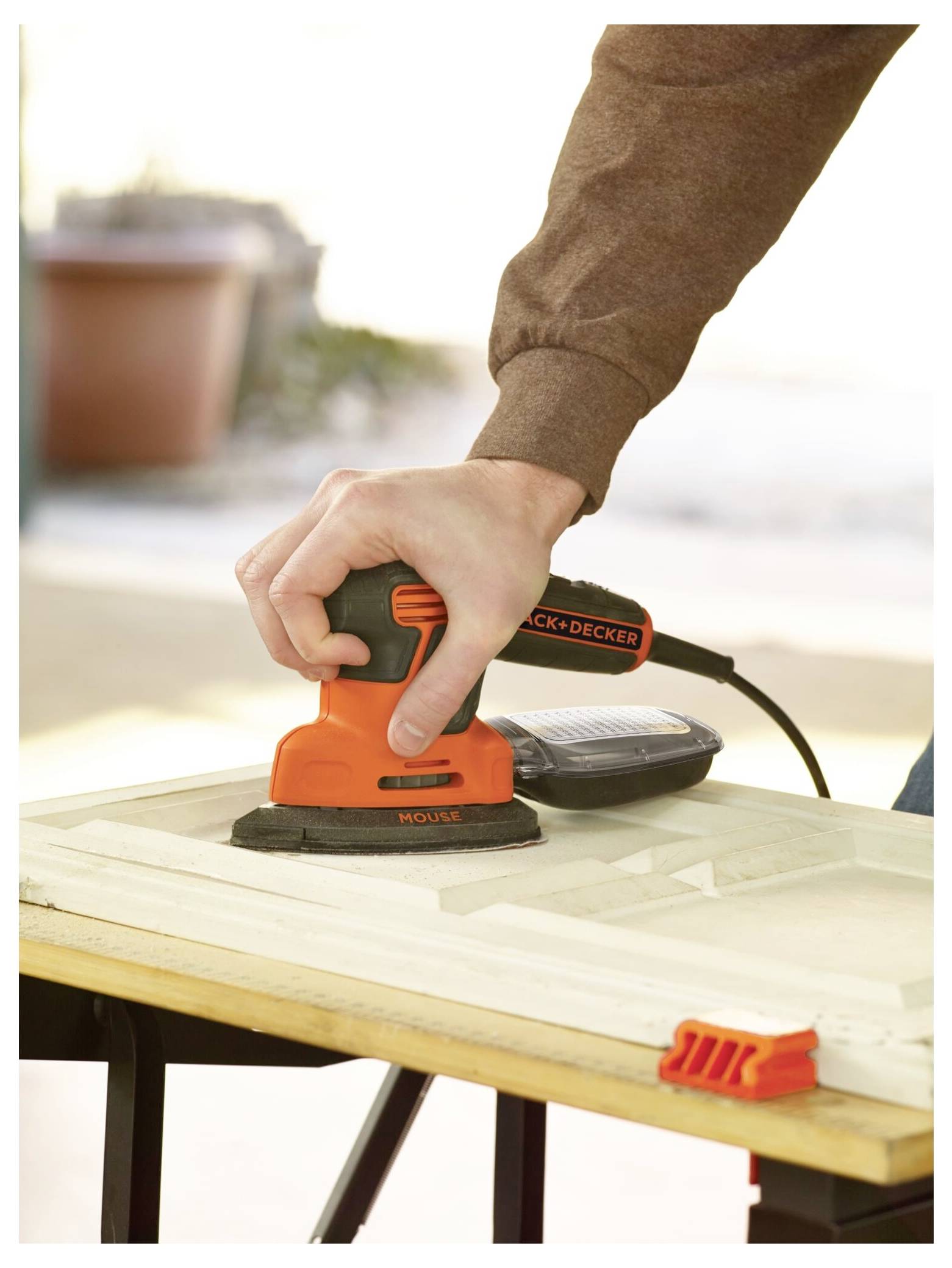 Eine Person verwendet einen orangefarbenen Black+Decker-Elektroschleiffer auf einer Holzoberfläche, mit Fokus auf den Griff der Hand und das Werkzeug in Betrieb.