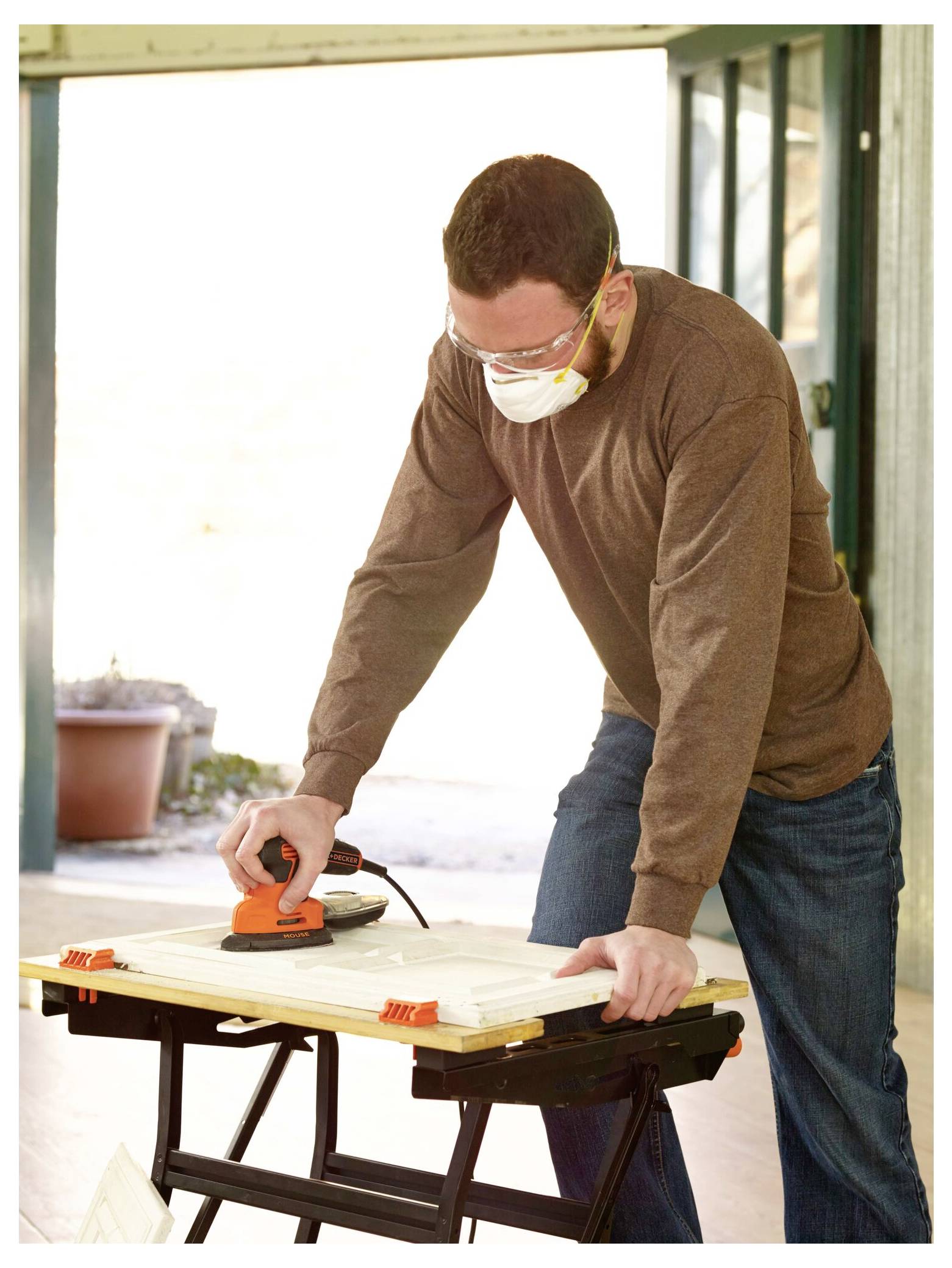 Eine Person, die eine Schutzbrille und eine Maske trägt, verwendet einen Elektroschleiffer auf einer Holzplatte, die auf einem Sägebock in einer gut beleuchteten Werkstatt positioniert ist.