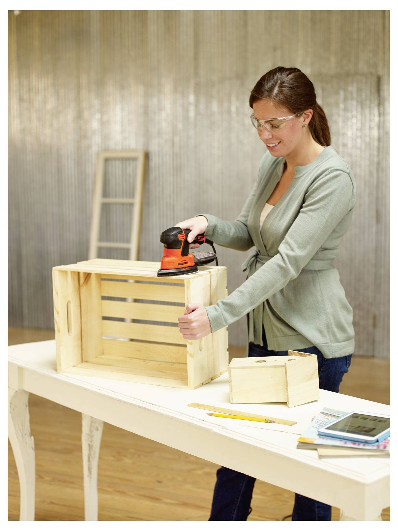 Eine Person verwendet einen tragbaren Elektroschleiffer zum Glätten einer Holzkiste, während sie an einem Tisch mit Werkzeugen und Materialien steht. Eine Schutzbrille wird getragen.