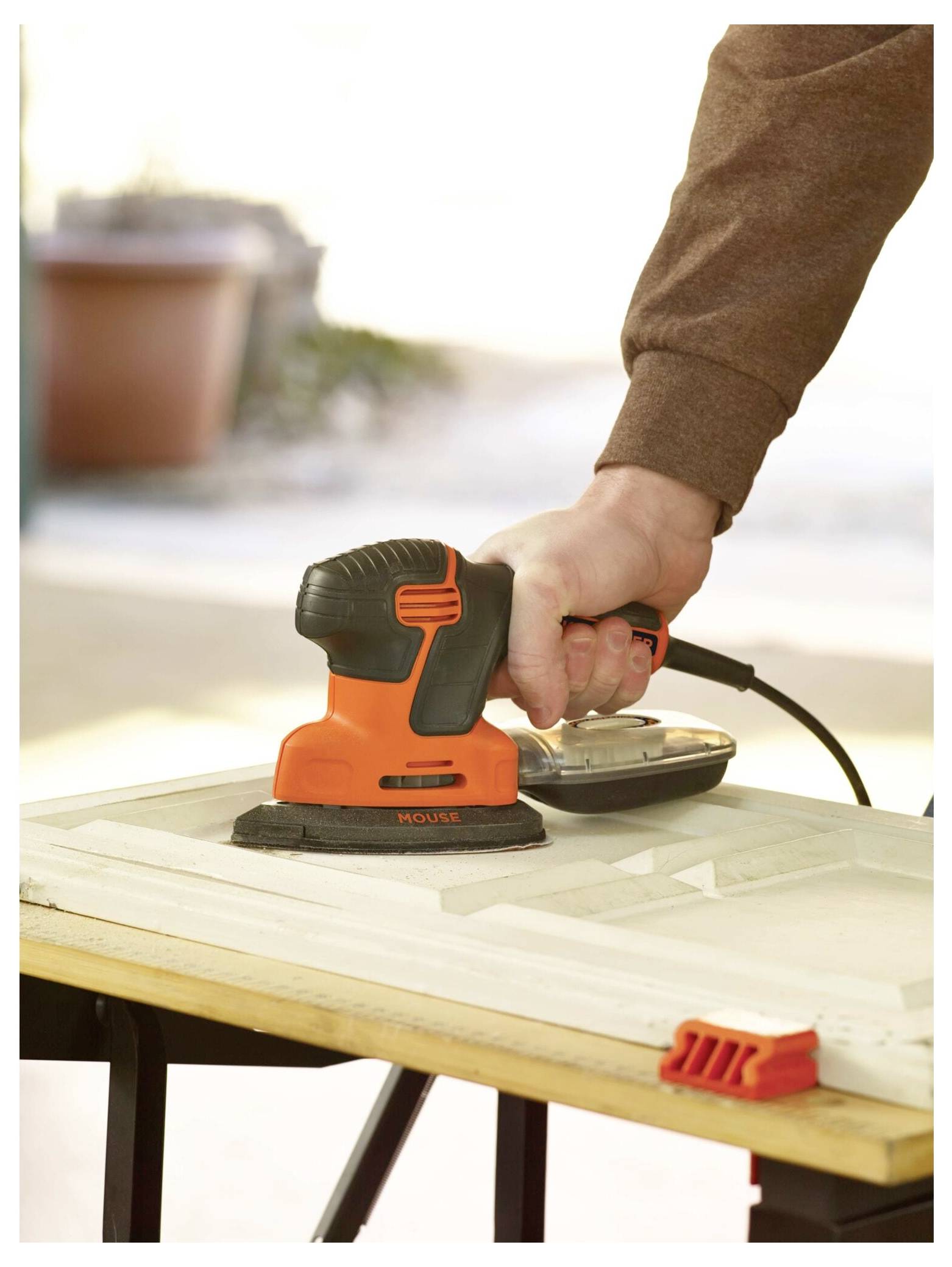 Eine Person schleift eine Holzoberfläche mit einem orange-schwarzen elektrischen Schleifergerät ab, draußen in einem sonnigen Bereich mit unscharfem Hintergrund.