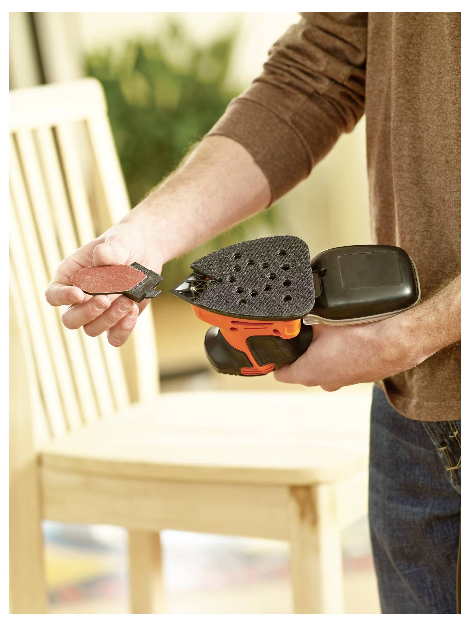 Eine Person demonstriert das Anbringen eines Schleifpapiers an einen tragbaren Elektroschleiffer, mit einem Holzstuhl im Hintergrund.