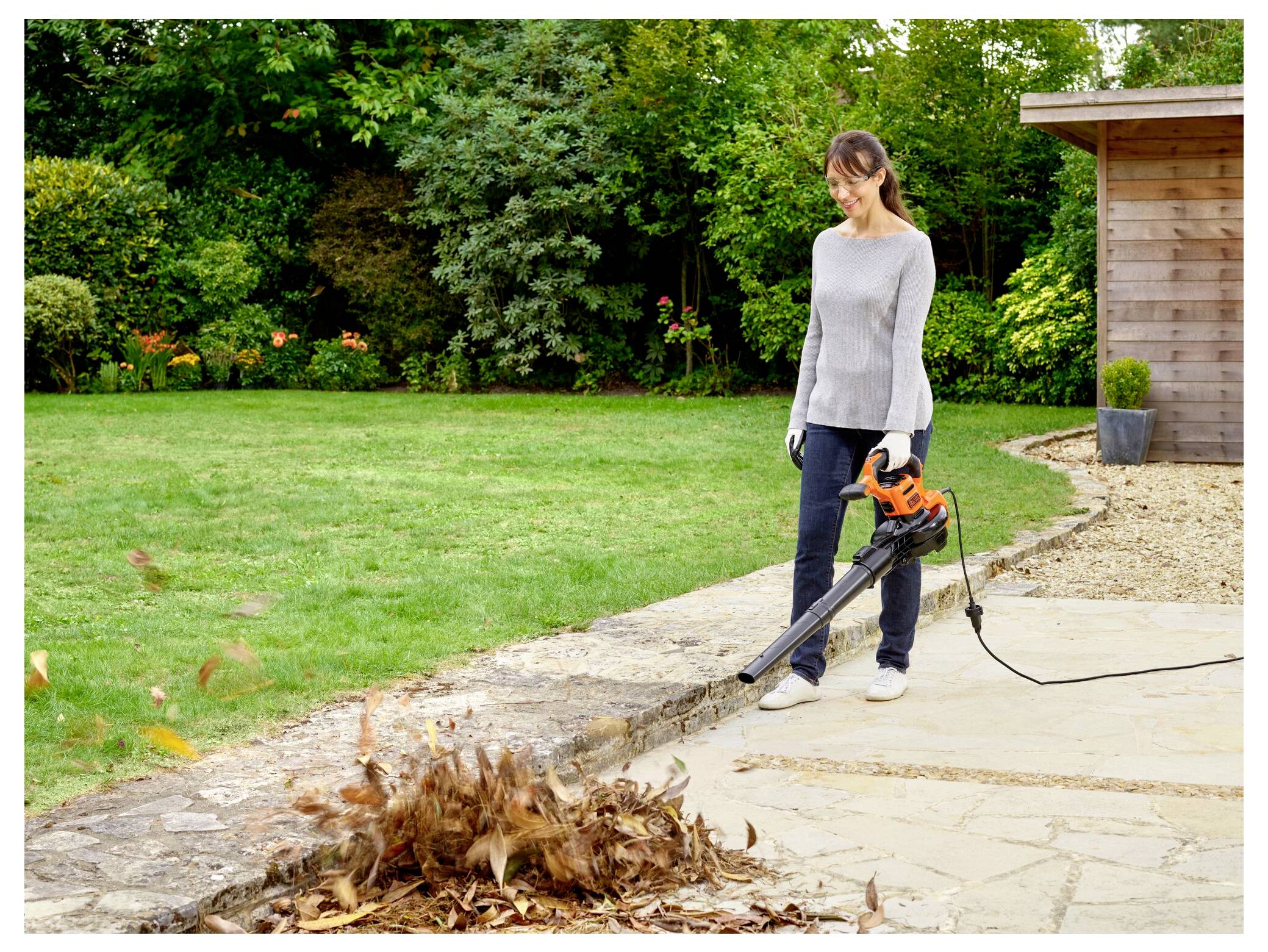Eine Frau, die ein Laubgebläse benutzt, um Blätter von einer Terrasse neben einem Garten zu entfernen. Sie trägt Freizeitkleidung und Handschuhe und schaut auf einen Holzschuppen.