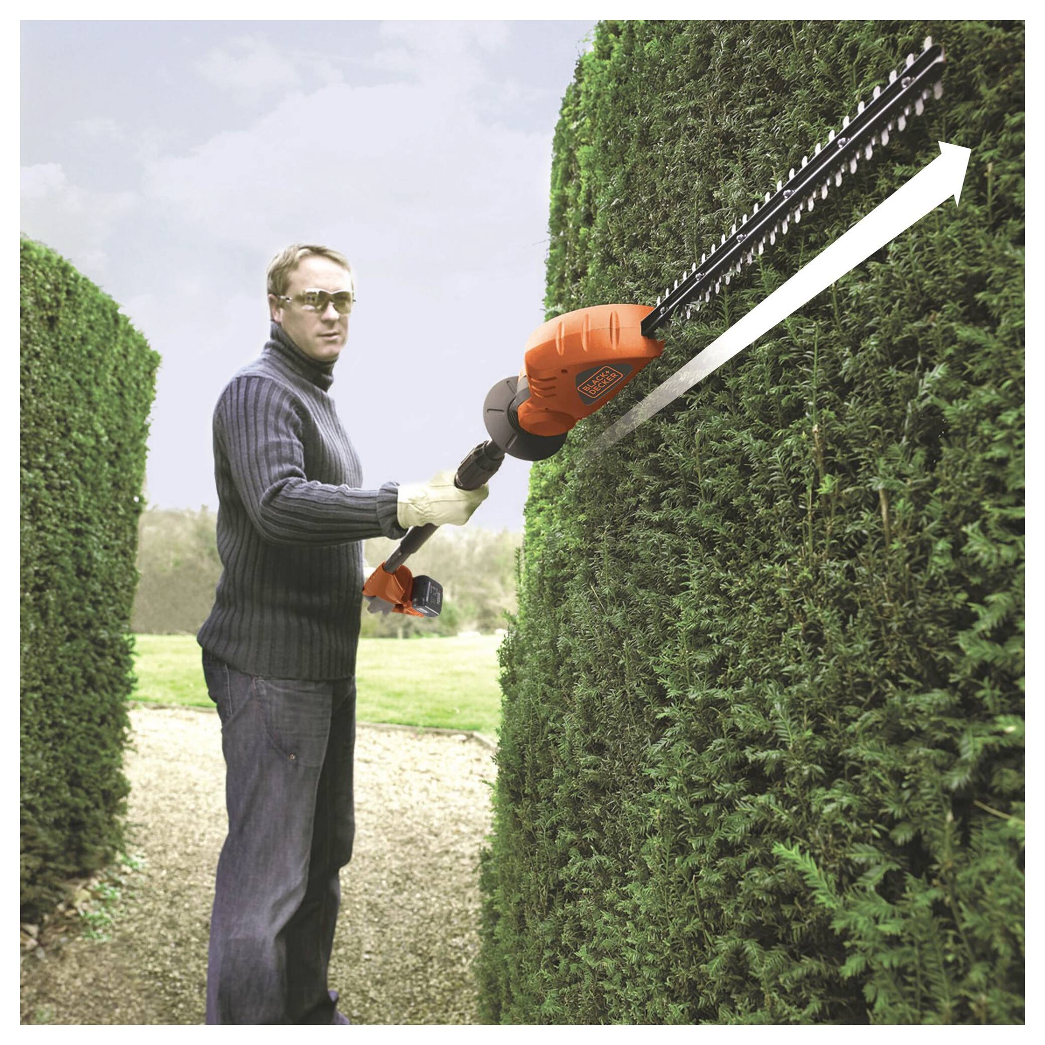 Eine Person, die eine Sonnenbrille und einen Pullover trägt, benutzt einen orangefarbenen Elektro-Heckenschneider, um hohe, grüne Hecken in einem Garten ordentlich zu schneiden.