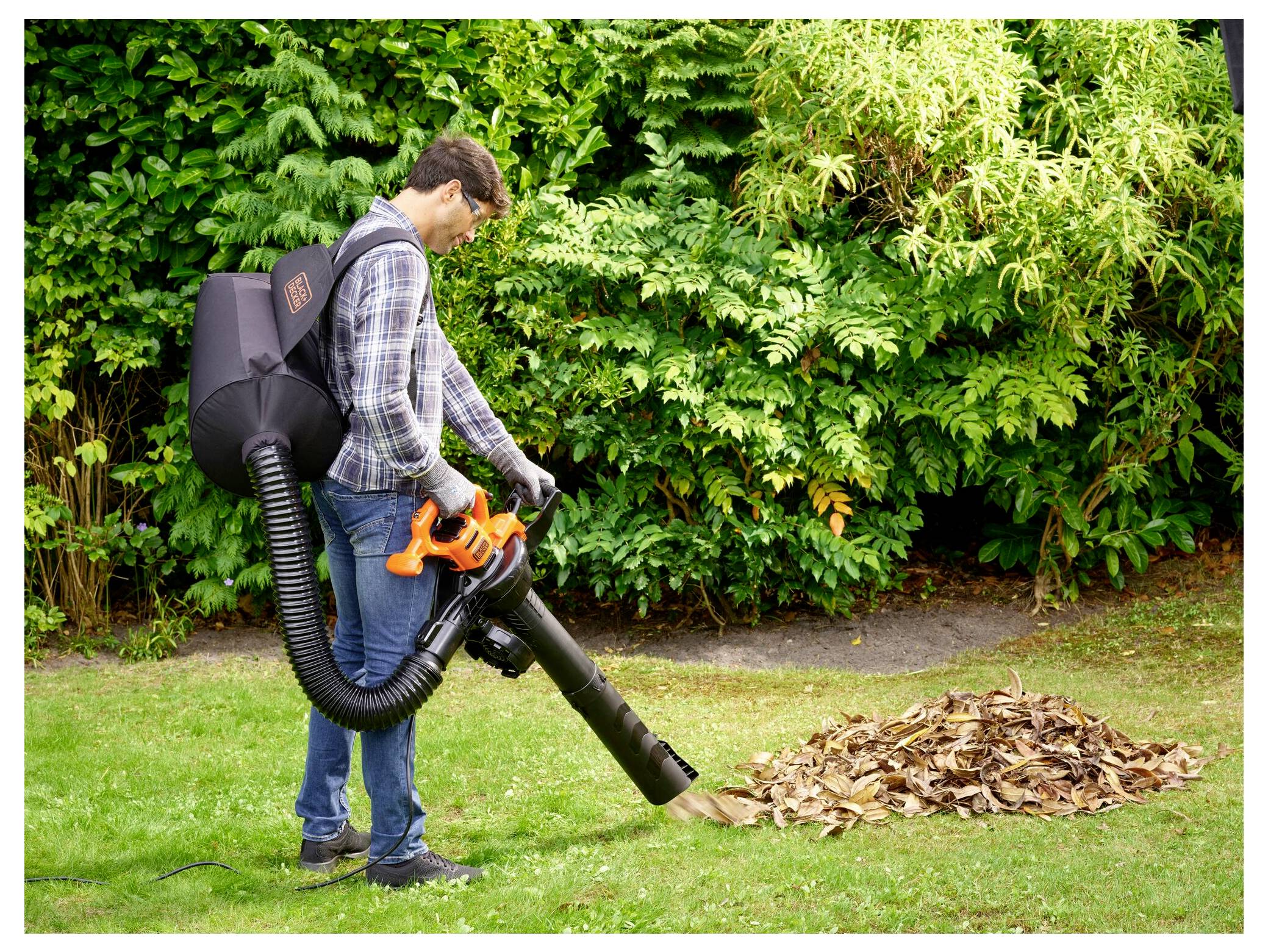 Eine Person benutzt einen Laubblaser/Staubsauger in einem Garten und räumt einen Laubhaufen auf einer Grünfläche auf, mit grünen Büschen im Hintergrund.