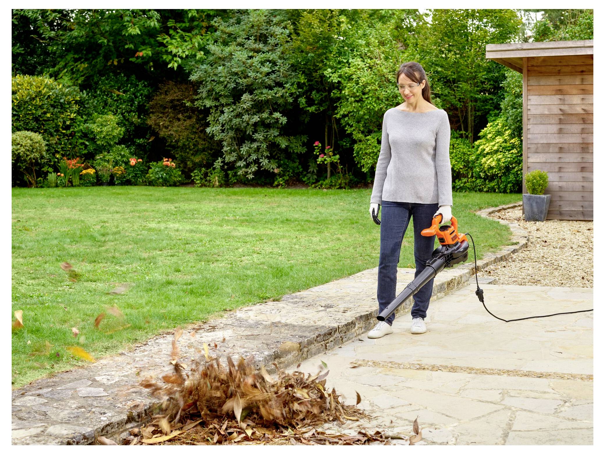 Eine Frau benutzt ein Laubgebläse auf einer Steinterrasse und bläst Blätter zu einem ordentlichen Haufen zusammen. Im Hintergrund sind ein Holzschuppen und ein Garten mit Büschen zu sehen.