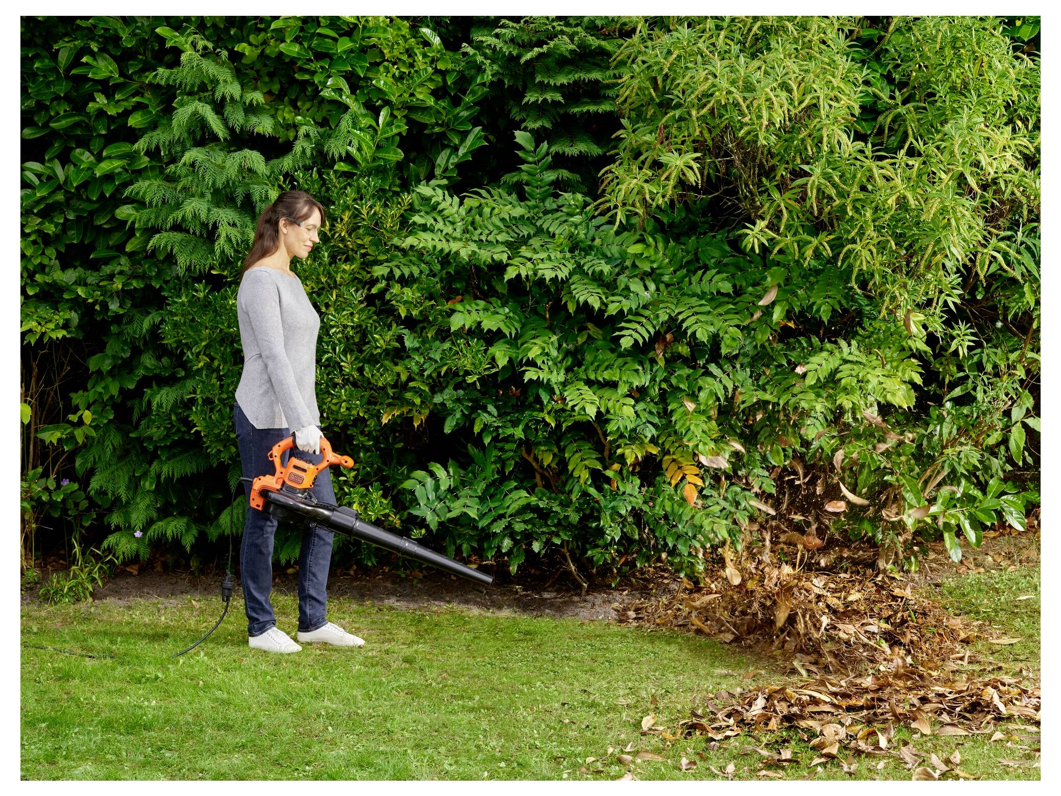 Eine Person benutzt ein elektrisches Laubgebläse, um gefallene Blätter auf einer grasbewachsenen Fläche zusammenzutragen, während dichtes grünes Laub im Hintergrund zu sehen ist.
