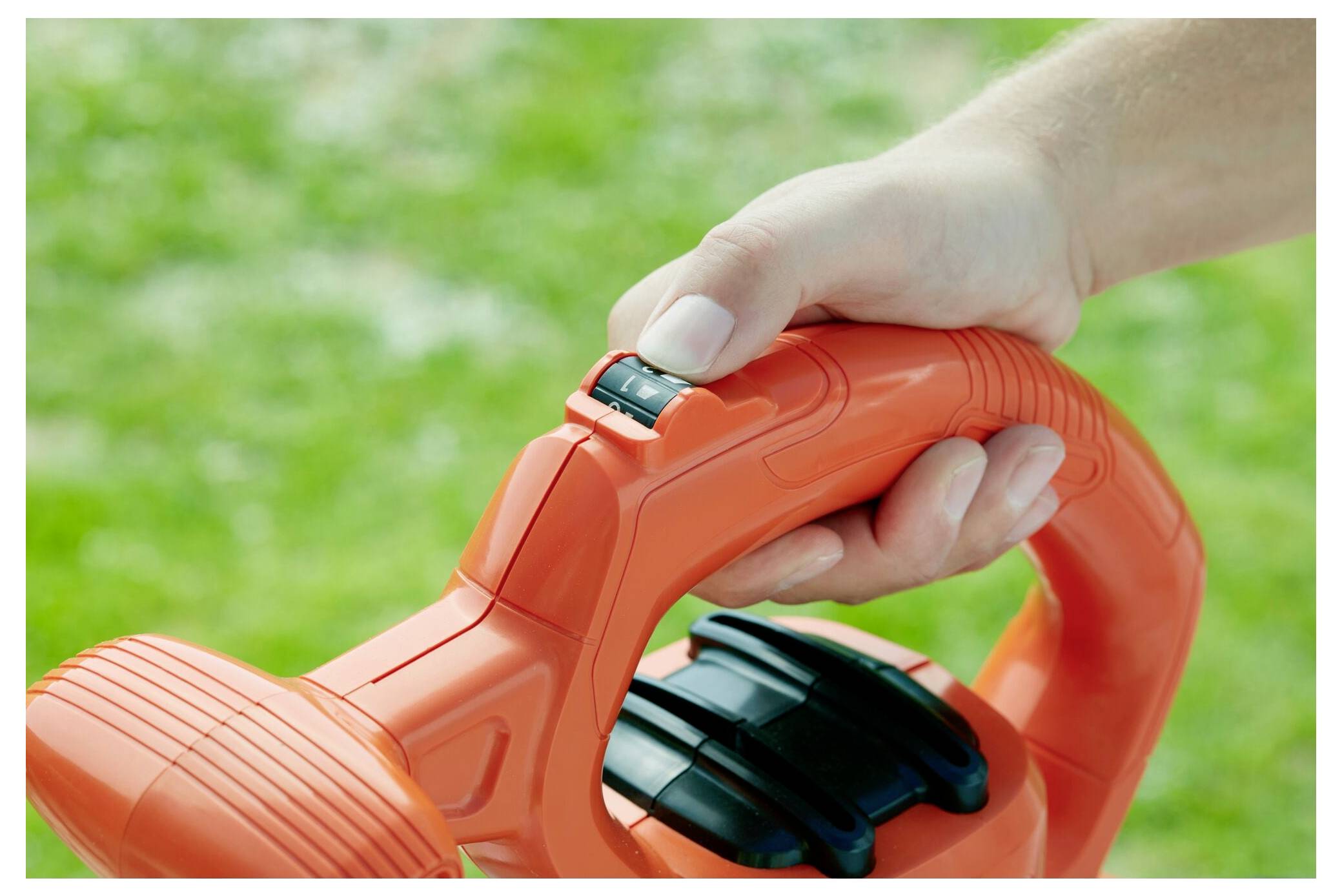 Eine Hand stellt die Geschwindigkeitseinstellung an einem orangefarbenen Gartengerät ein, mit einem grasbewachsenen Hintergrund.