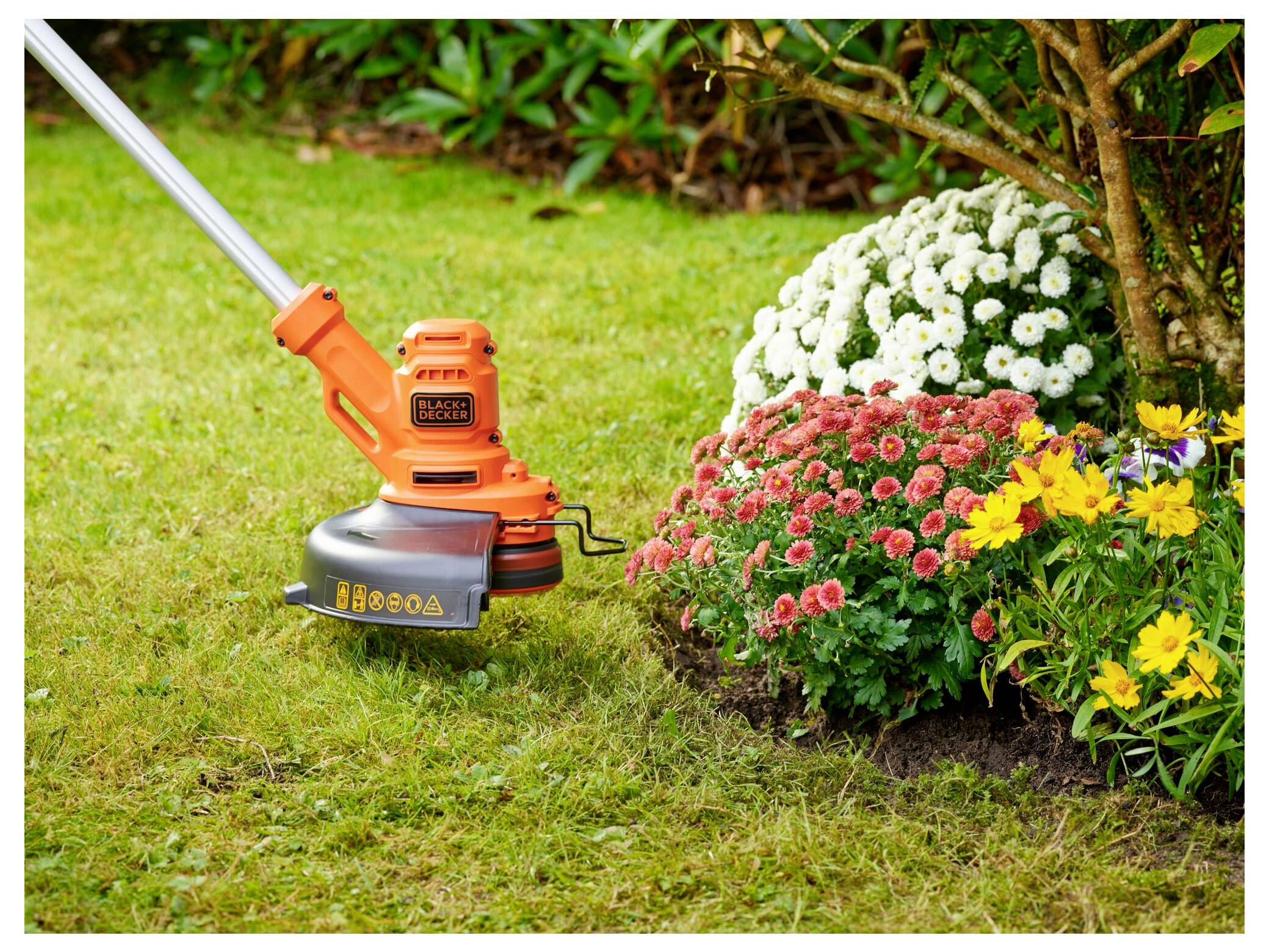 Orange-farbener elektrischer Fadentrimmer schneidet den Rasenrand neben einem Blumenbeet mit weißen, rosa und gelben Blüten in einer Gartenanlage.