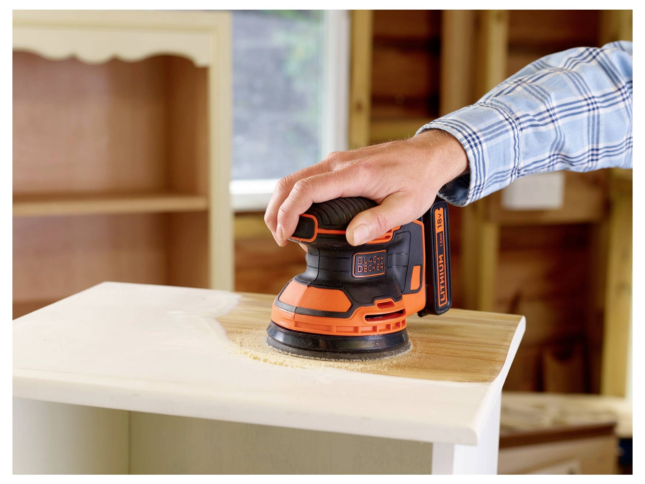 Eine Person benutzt einen orange-schwarzen elektrischen Elektroschleiffer, um die Oberfläche eines Möbelstücks aus Holz in einem Innenraum zu glätten.